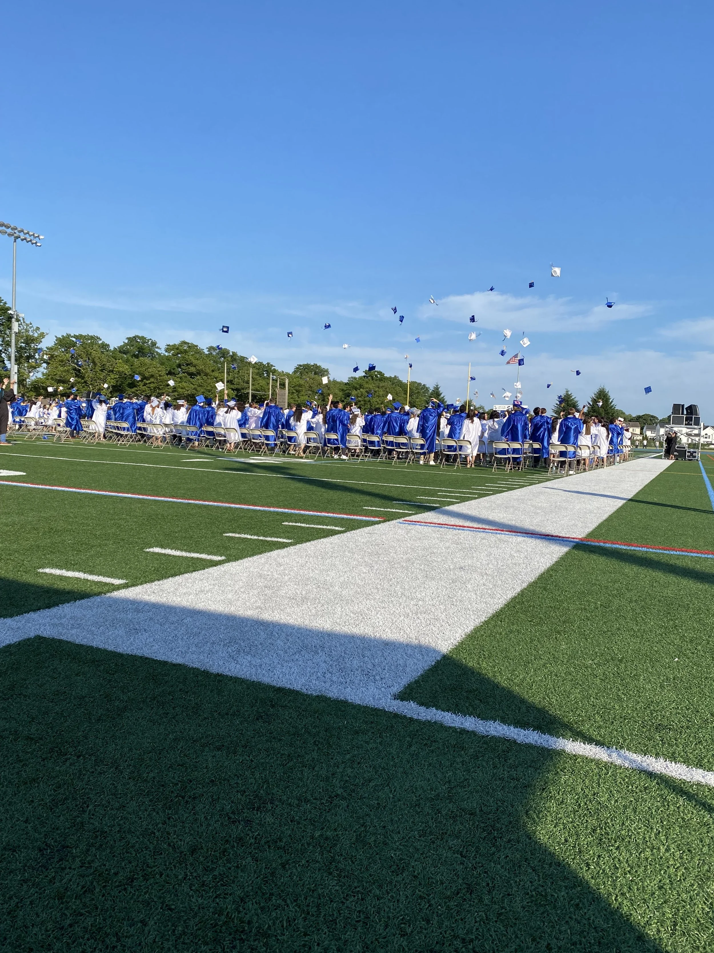 Graduates in blue and white caps and gowns during a graduation ceremony on a football field, with caps being tossed in the air under a clear blue sky.