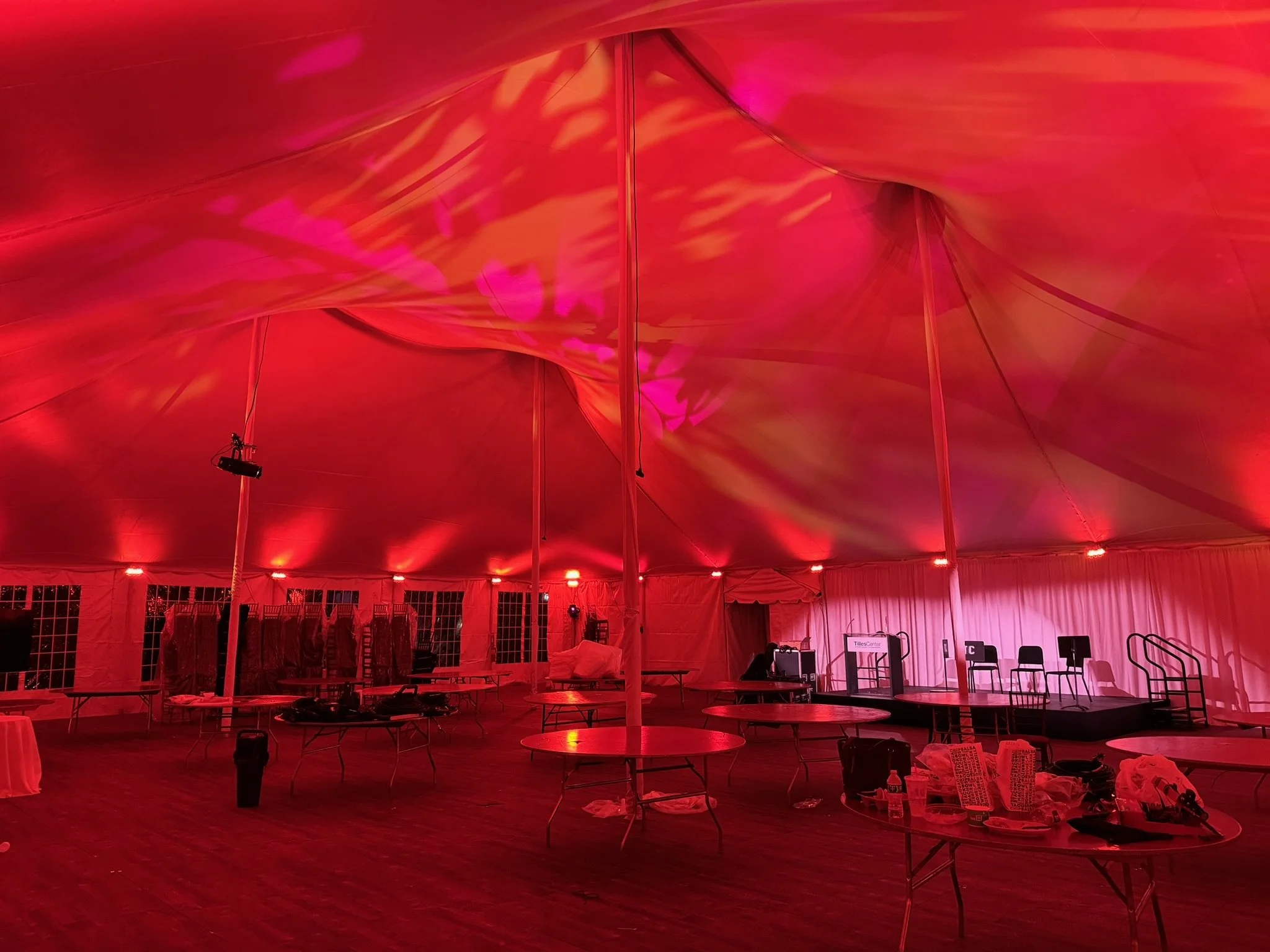 Interior of a large event tent with red lighting, tables, chairs, and stage setup.