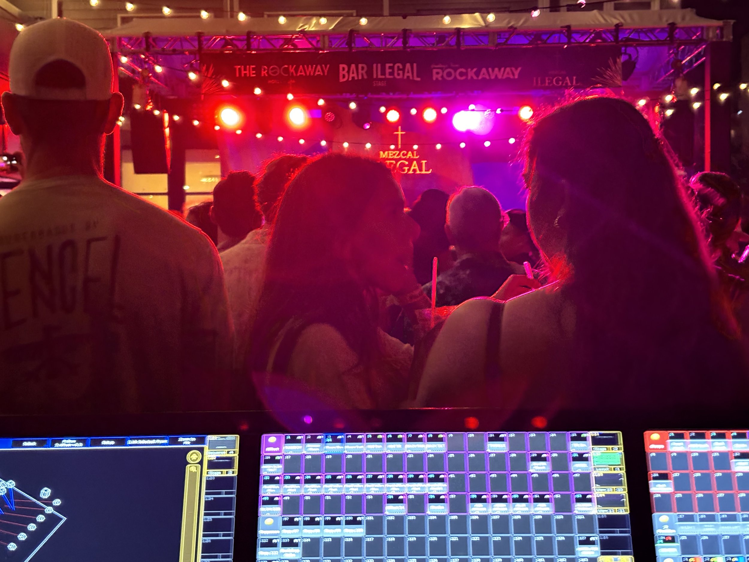 People sitting and watching a live music performance at an outdoor concert with stage lights and banners that read and 'Rockaway'. DJ equipment in the foreground.