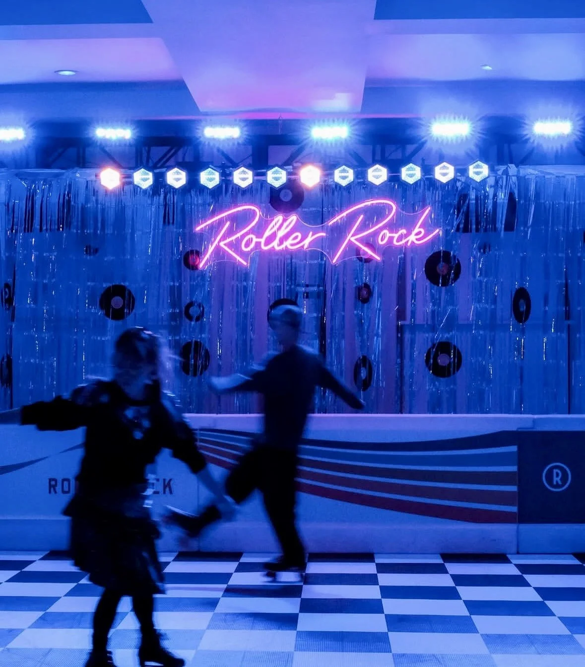 Two children roller skating in front of a neon sign that reads "Roller Rock" on a decorated stage with vinyl records and metallic streamers.