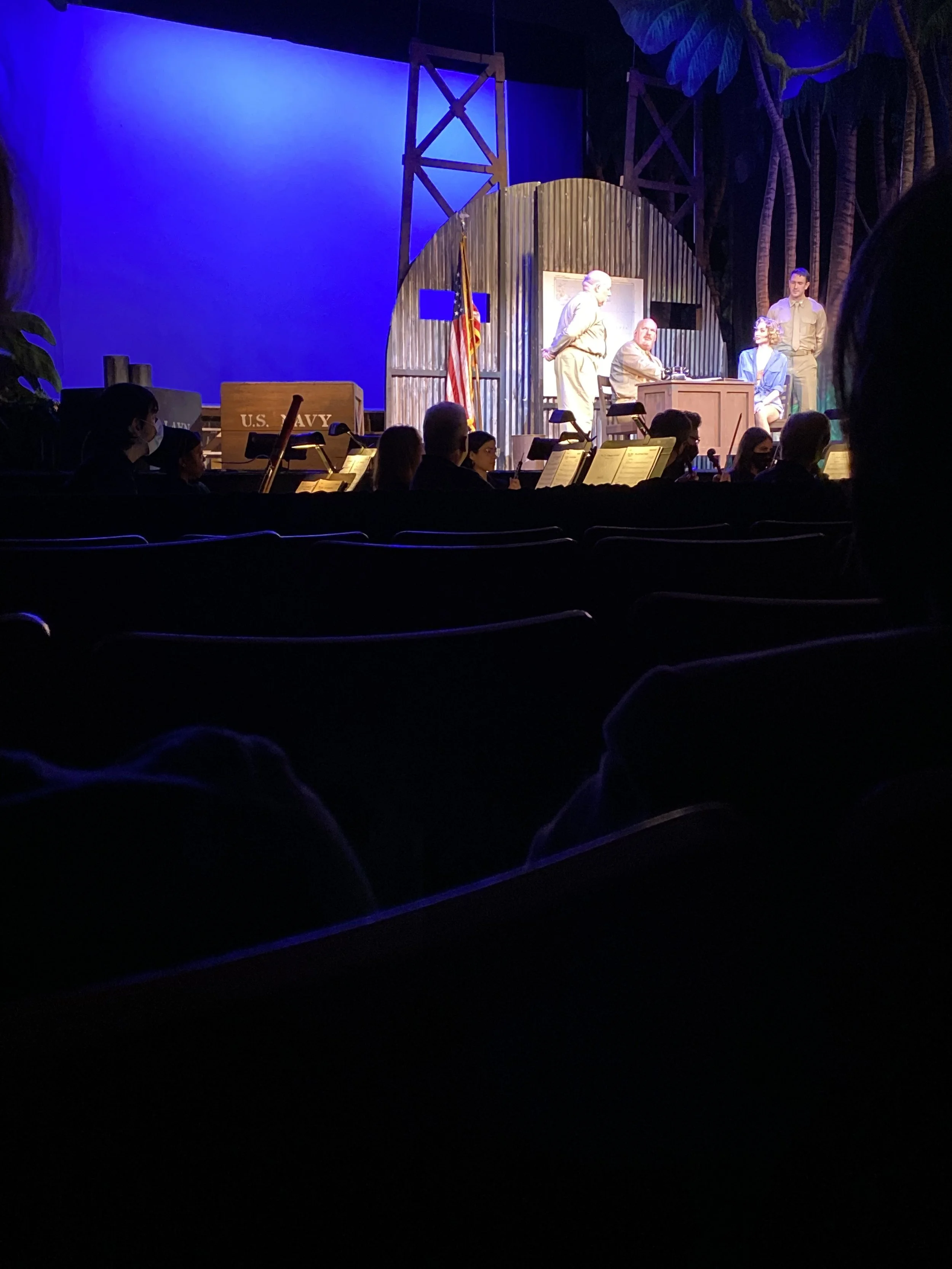 The image shows a theatrical stage with actors performing and an orchestra in the foreground. The stage is decorated with a U.S. Navy theme, including a box with 'U.S. Navy' written on it and an American flag. The theater audience is seated and watch