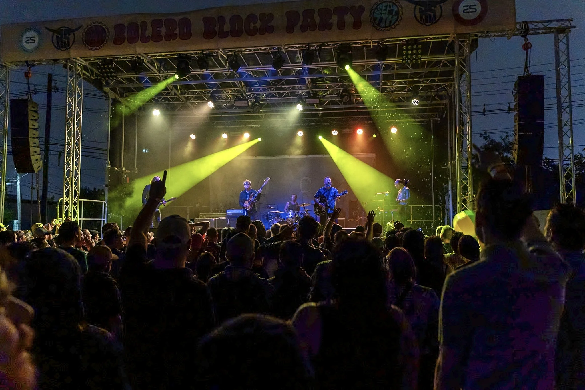 A live concert on an outdoor stage during evening with a band performing under yellow and red stage lights, and a crowd of people watching.