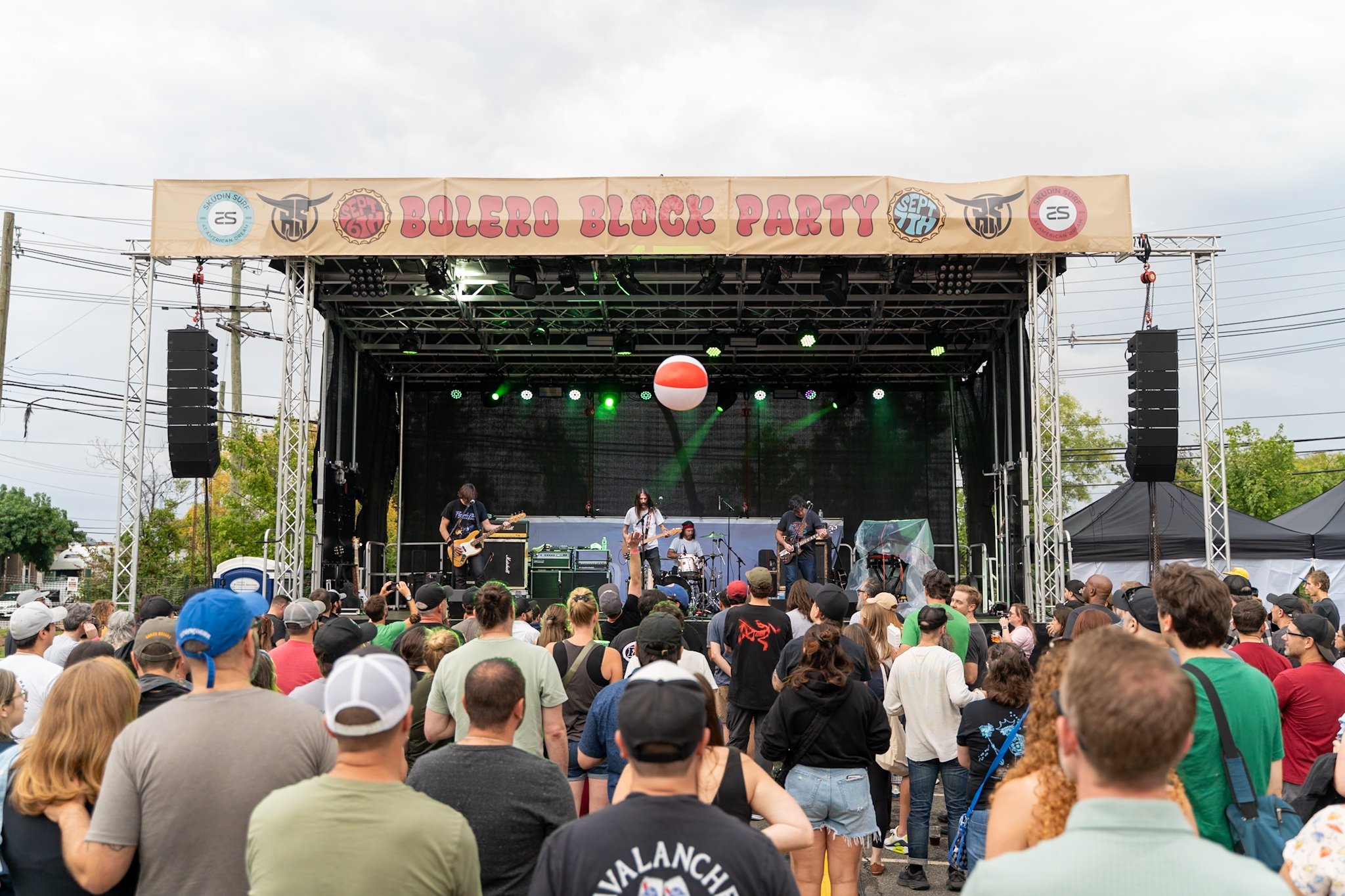 People watching a band perform on an outdoor stage at the Bolero Block Party, with a large crowd and a beach ball in the air.