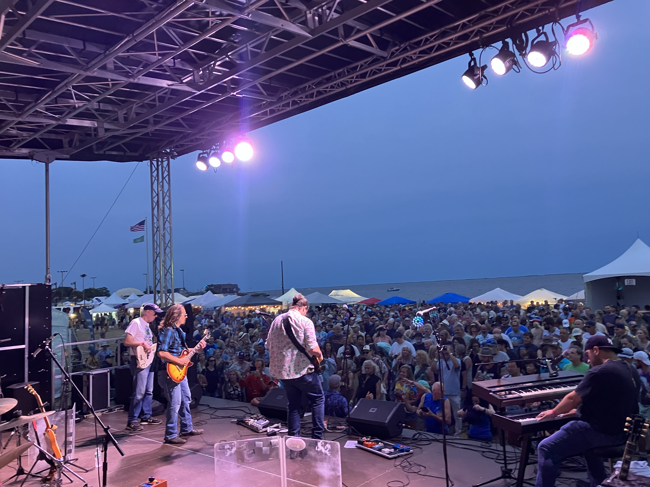 Live outdoor concert at dusk with a band performing on stage, audience gathered near the water, and tents in the background.