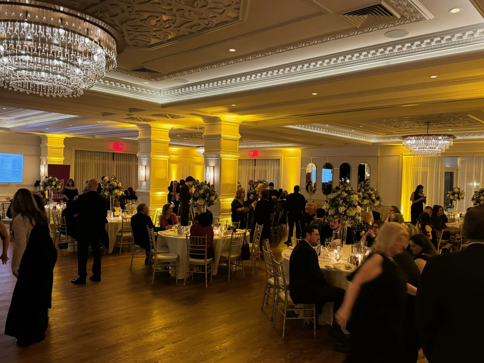 Elegant banquet hall decorated with chandeliers, large floral centerpieces, and yellow lighting, filled with people in formal attire seated at round tables and standing, engaged in conversation during an event or celebration.