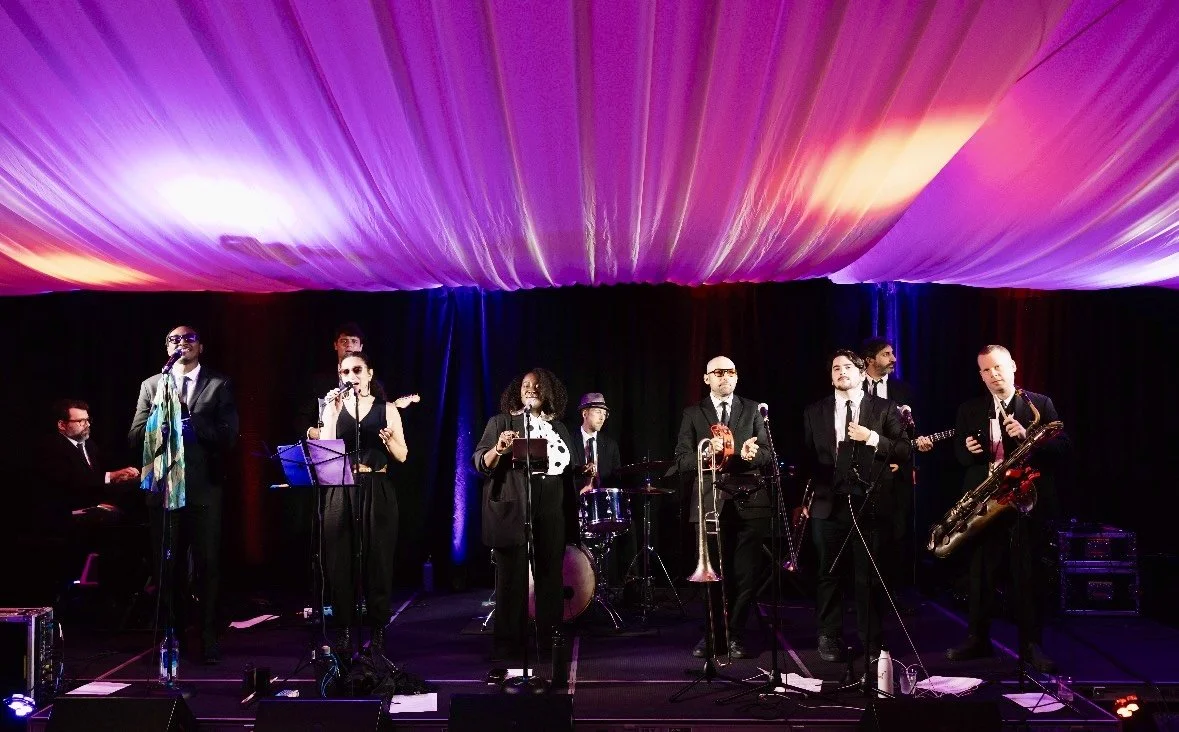 A jazz band performing on stage with seven musicians playing various instruments including keyboard, saxophone, trumpet, and drums, under a colorful, illuminated ceiling.