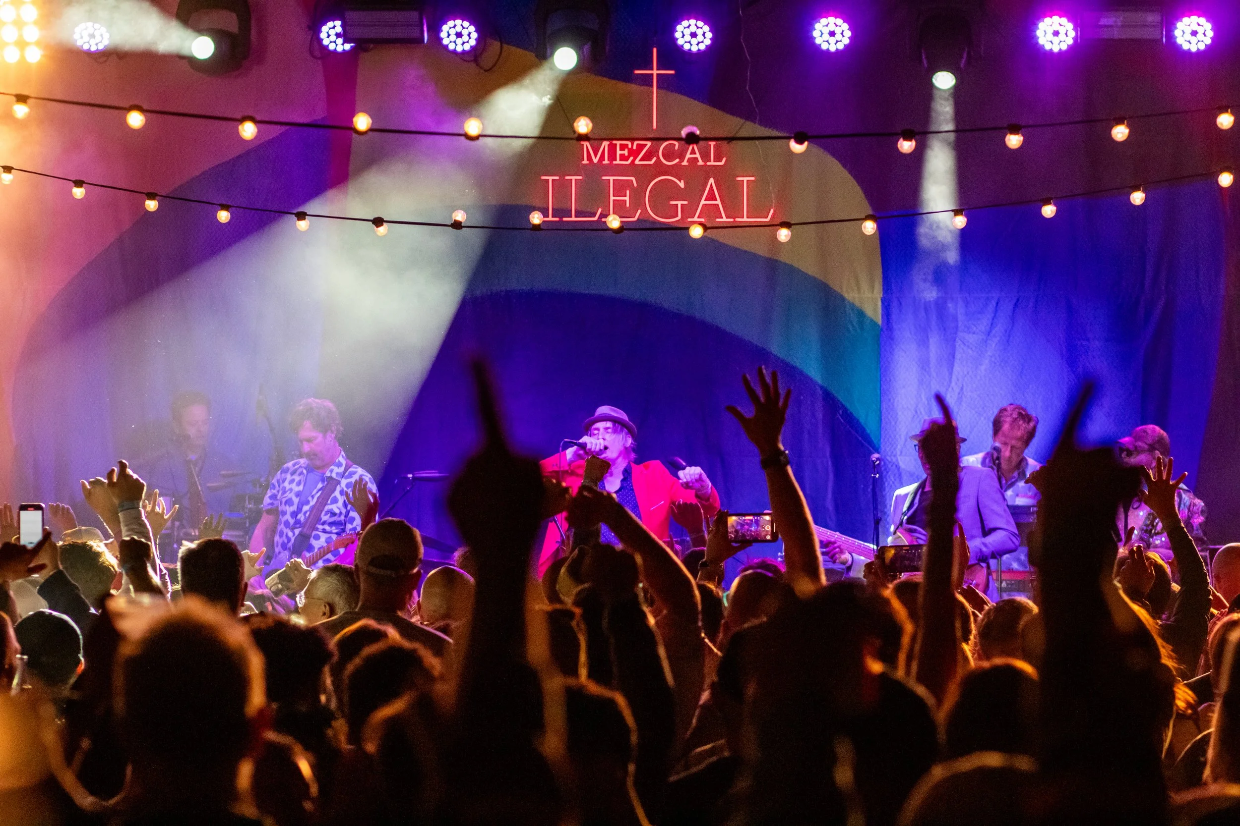 A live concert with a stage lit in colorful lights, featuring a band performing under a neon sign. The crowd in front of the stage is raising their hands and taking photos.
