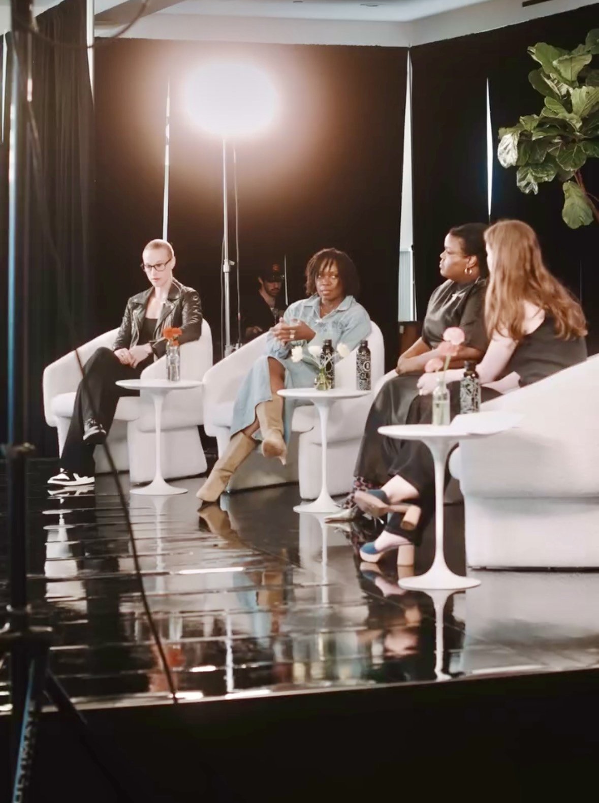 Four women sitting on white couches during a panel discussion, with small tables holding flowers and bottles in front of each woman, a bright studio light overhead, and a reflection on the shiny floor.