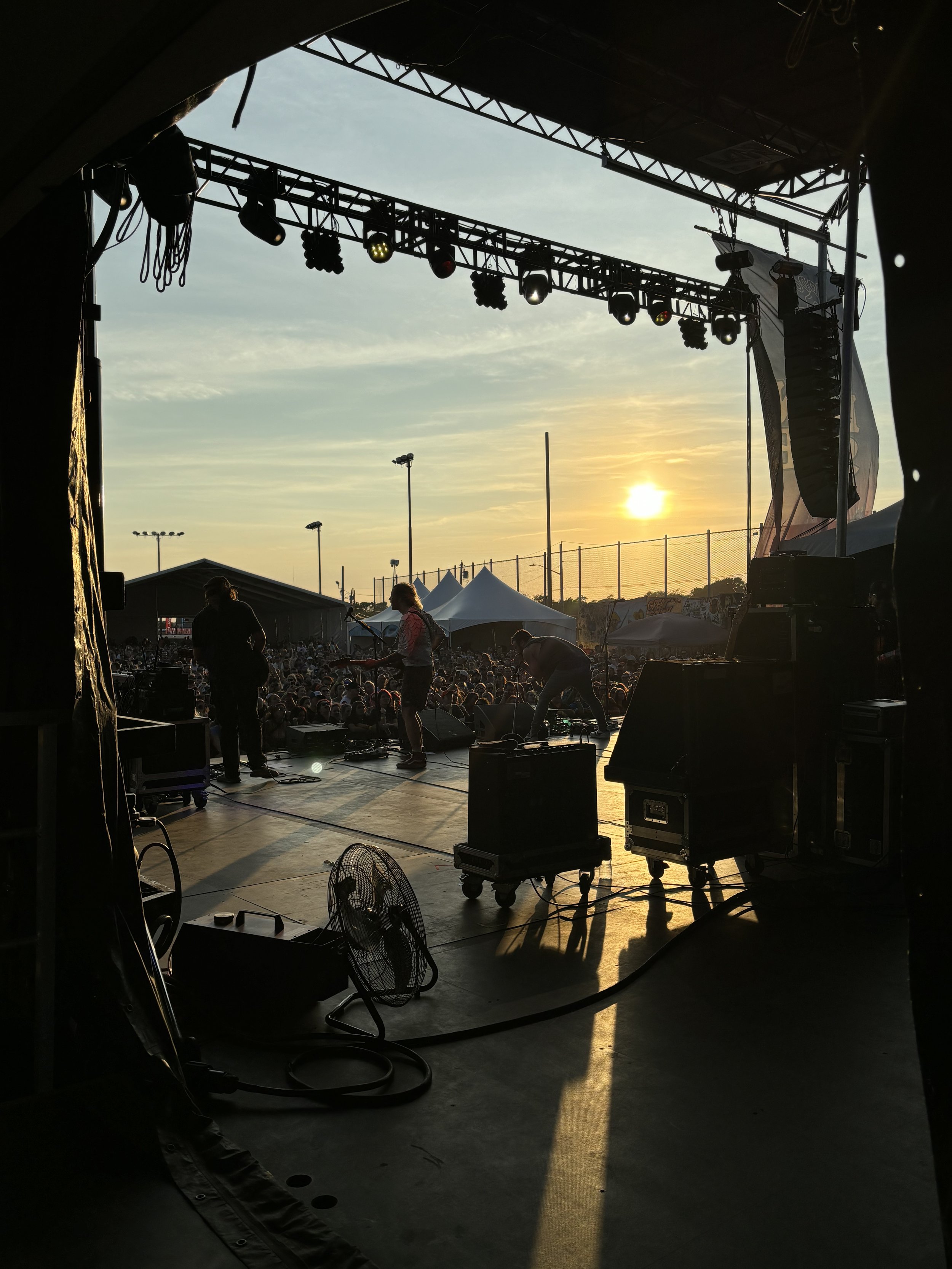 View from inside a concert stage looking out at a crowd at sunset. There are musicians on stage preparing for a performance, with equipment and gear visible.