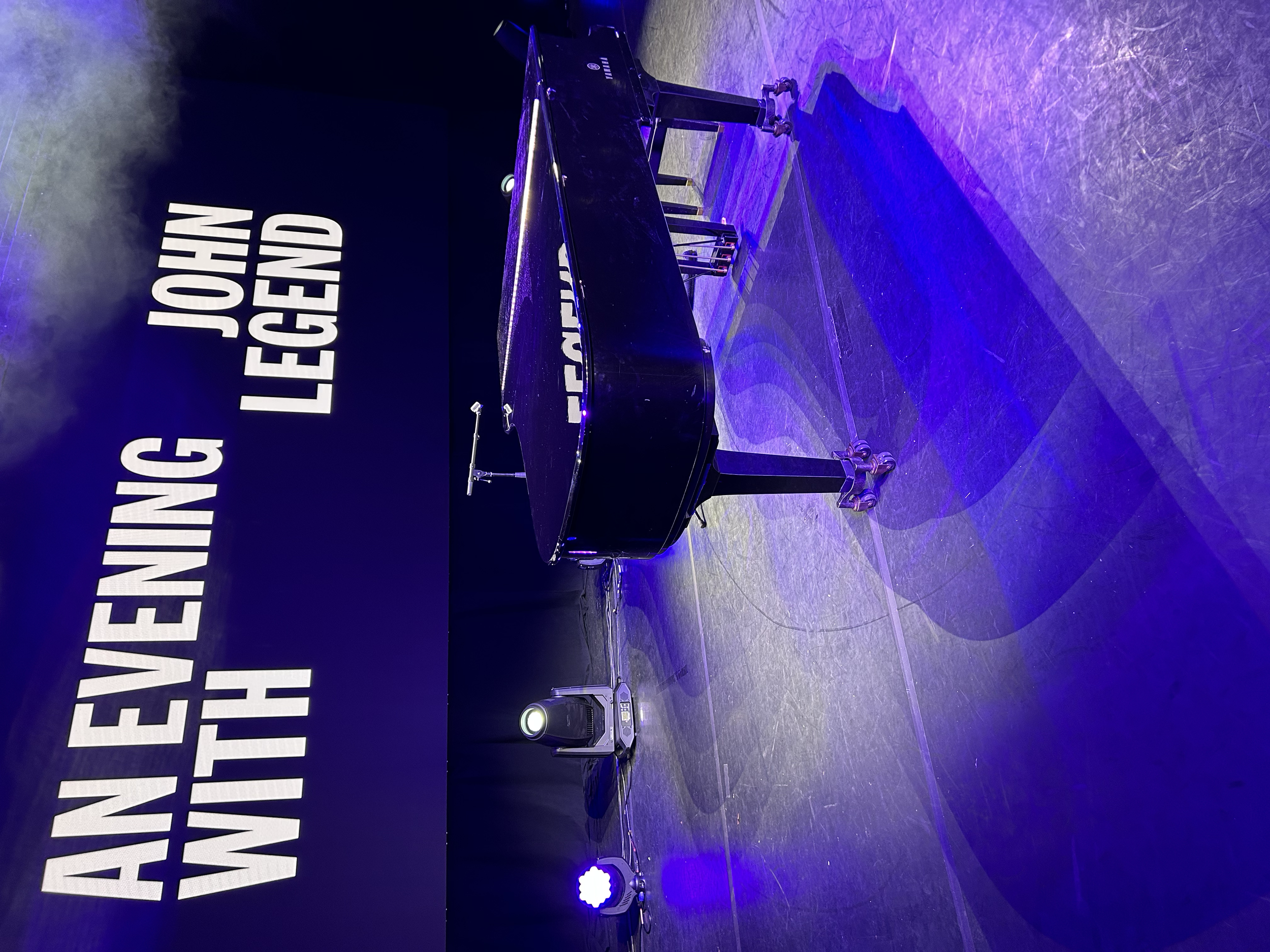 A grand piano set on a dark stage with blue and purple lighting. Behind it, there is a black wall with large white text that reads 'AN EVENING WITH JOSH LEGGEND.'