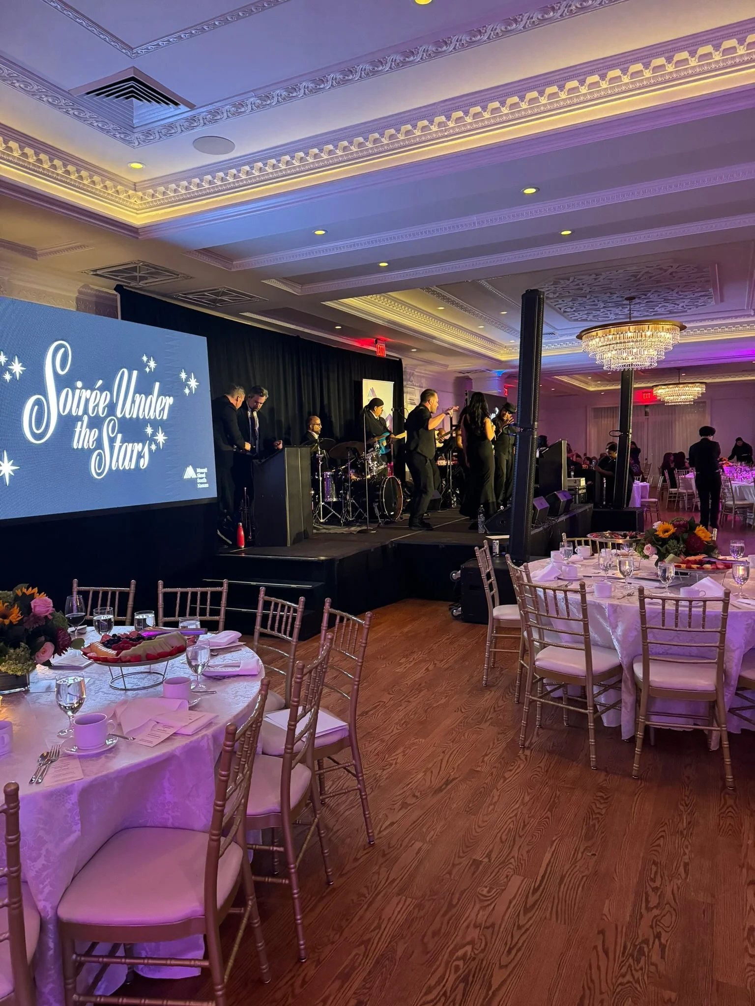 A band performing on stage at a formal event called 'Soirée Under the Stars' in a decorated banquet hall with round tables, floral centerpieces, and elegant place settings.