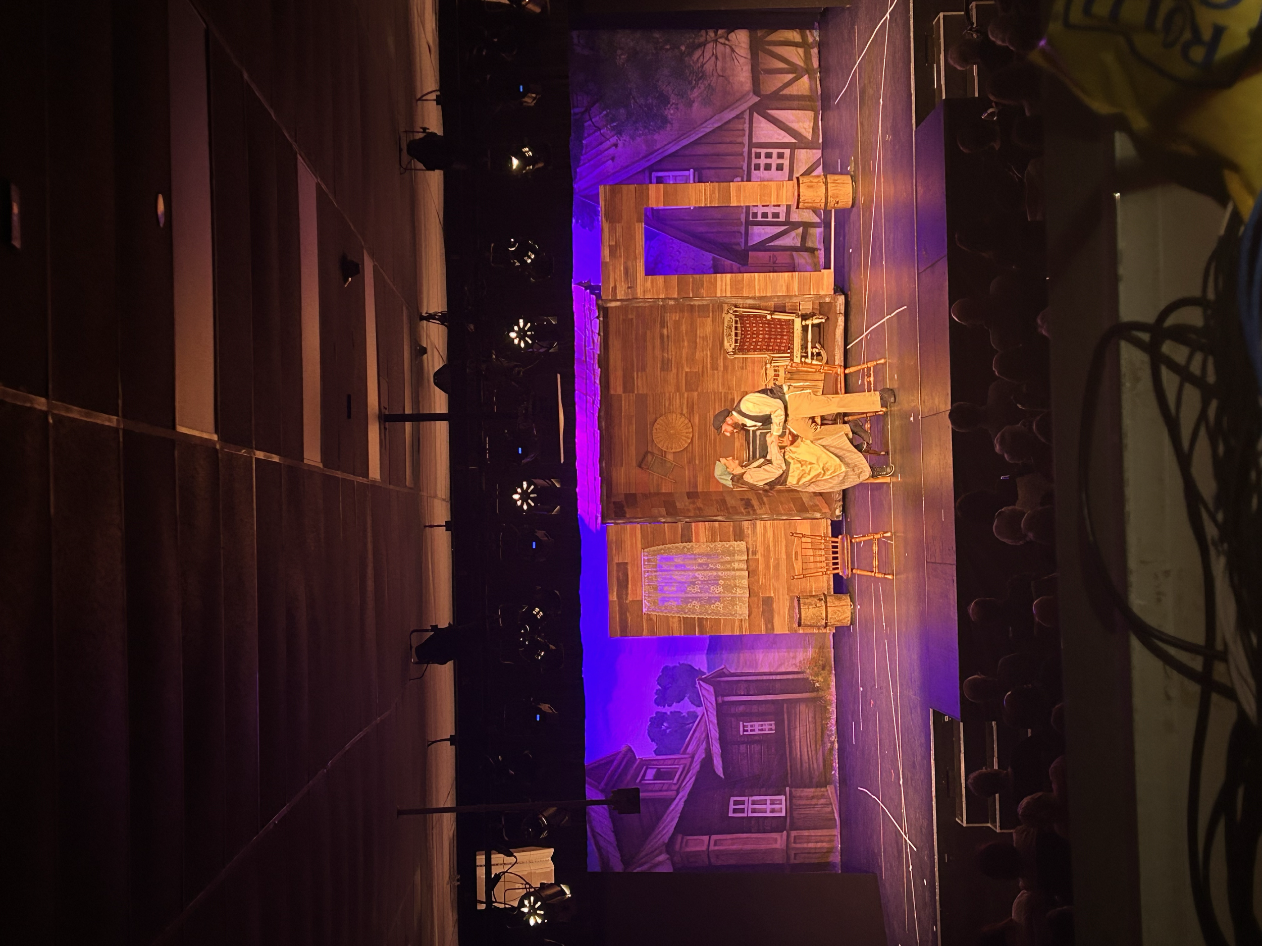 A stage set with wooden walls and furniture, depicting a rustic room with a small house and garden, and two performers dressed in period costumes engaged in a theatrical scene, with a backdrop of a farm and a sunset sky.