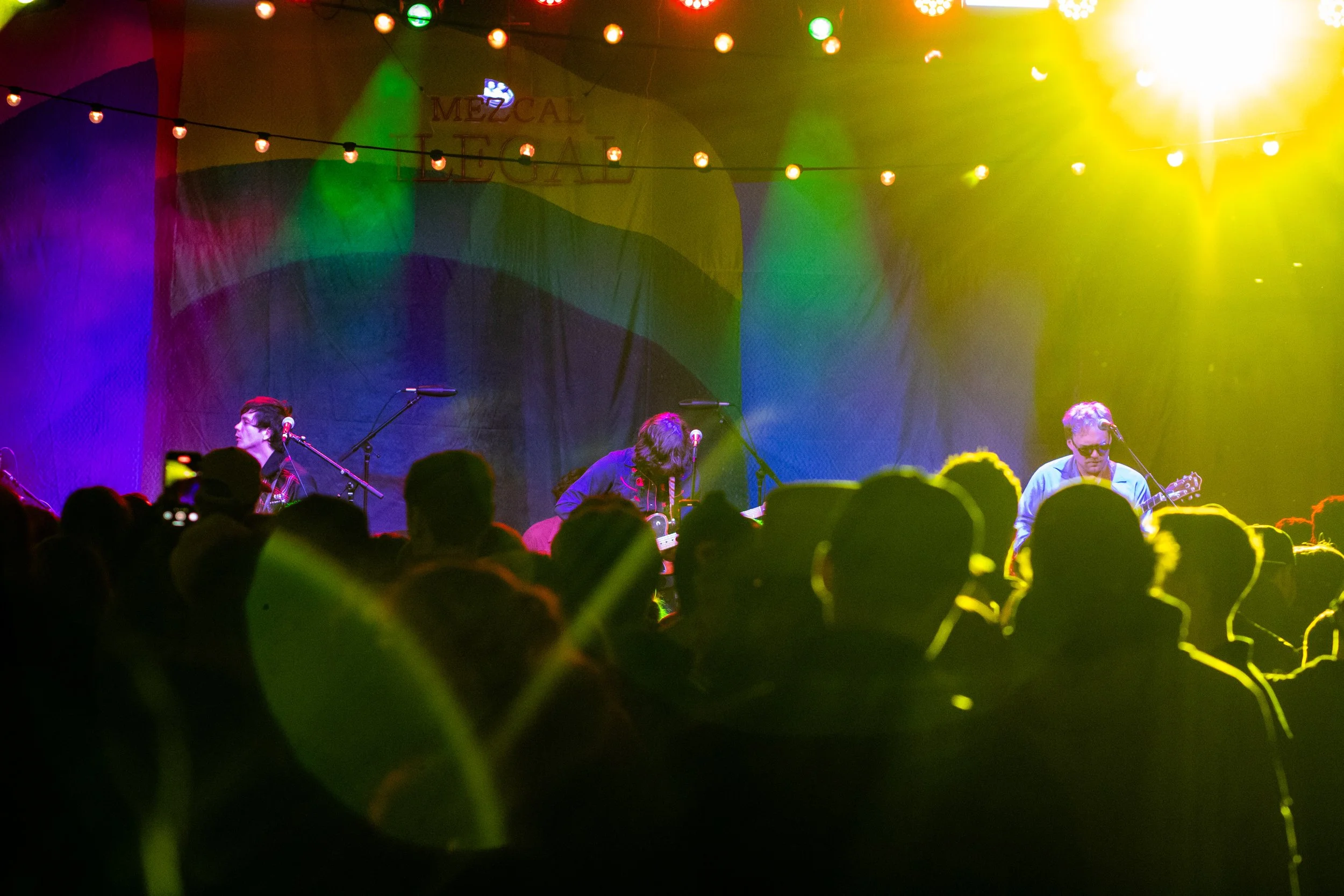 A band performs on stage with colorful lighting and an audience watching in the foreground.