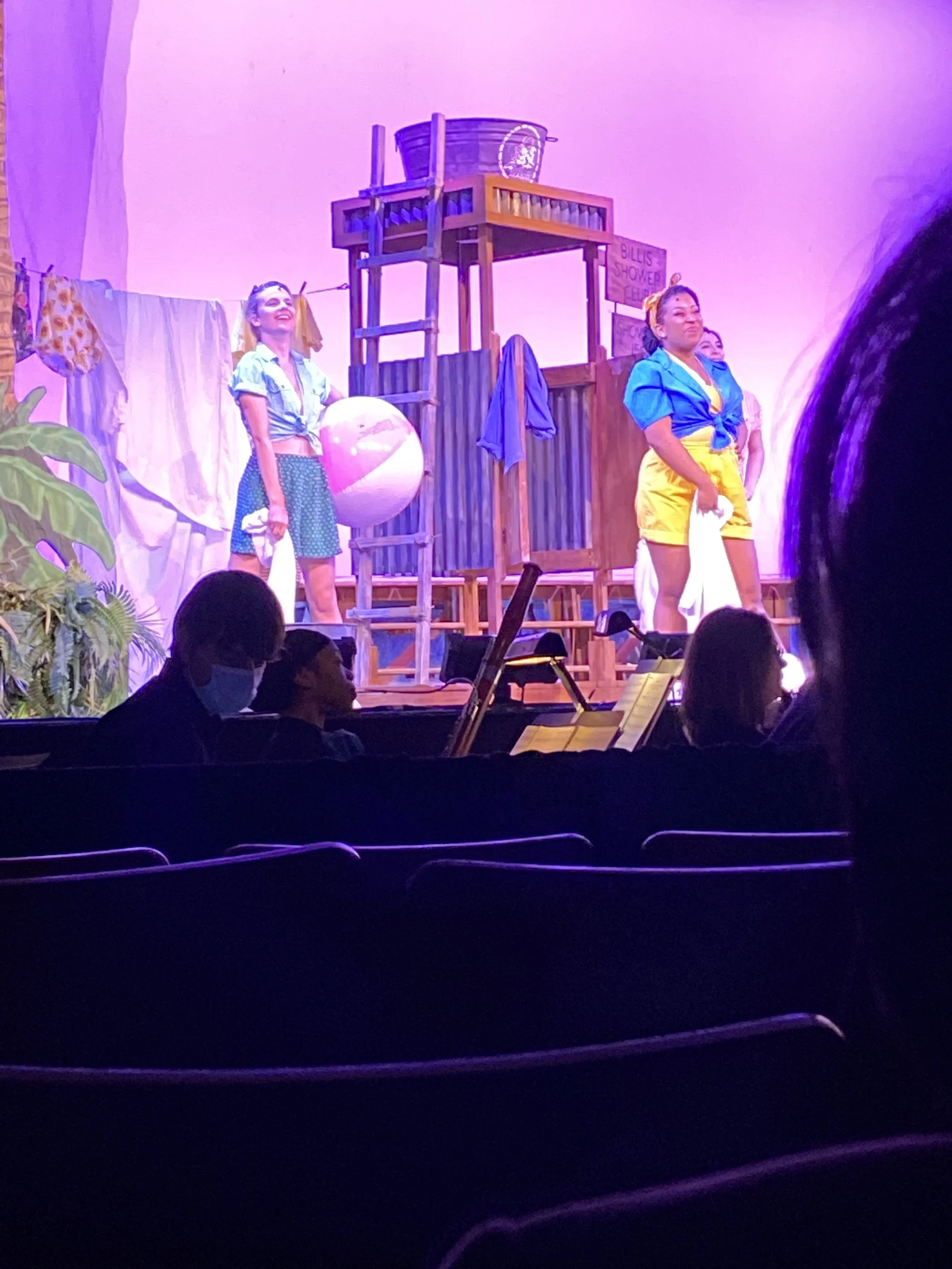 Stage scene from a play with actors in colorful costumes, set like a beach with props like a beach ball, ladder, and shower sign, with audience members watching.