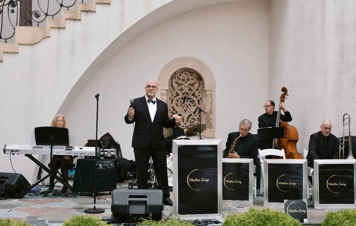 A jazz band performing outdoors, with a man in a tuxedo singing into a microphone, accompanied by musicians playing keyboard, drums, saxophone, upright bass, and trombone.