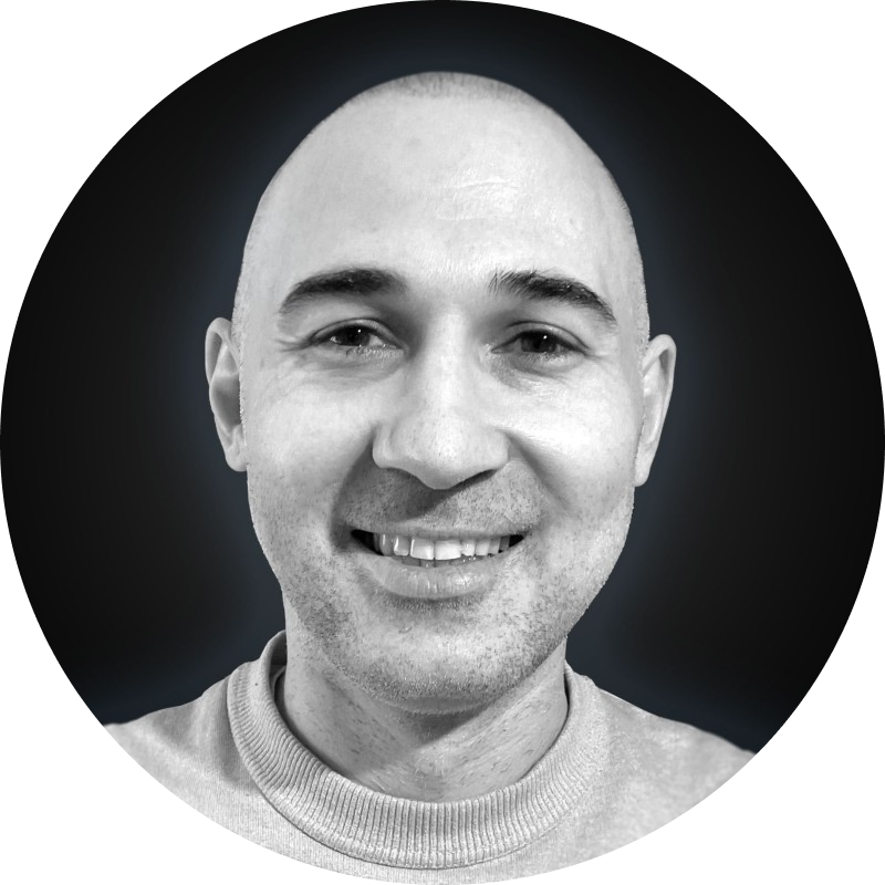 Black and white close-up photo of a smiling man with a shaved head wearing a light-colored crewneck sweater.