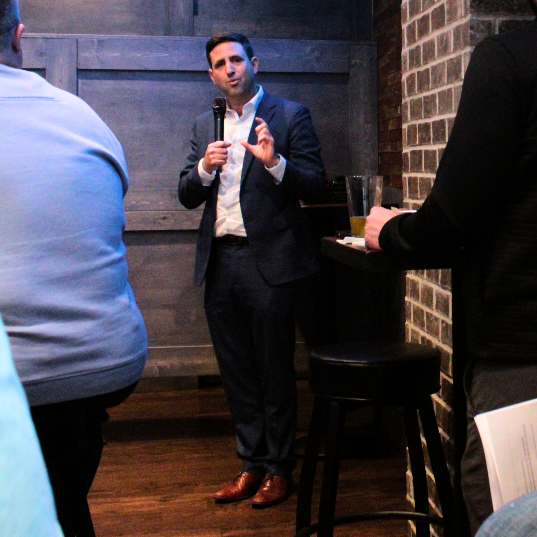 Man in a dark suit and white shirt giving a speech or presentation, holding a microphone, in a dimly lit venue with brick and wood wall decor.