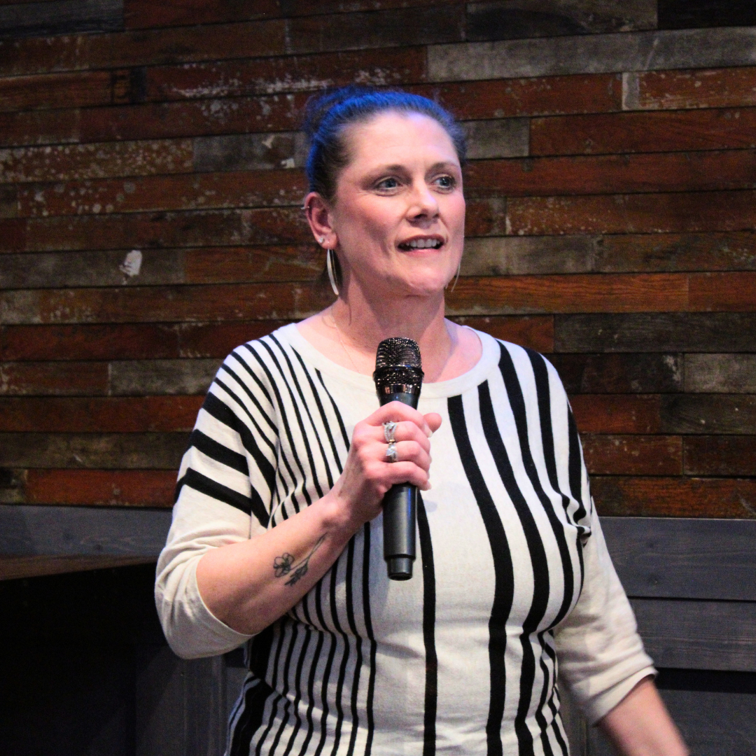 A woman with dark hair in a bun, wearing a black and white striped shirt, holding a microphone and speaking, standing against a wooden wall background.