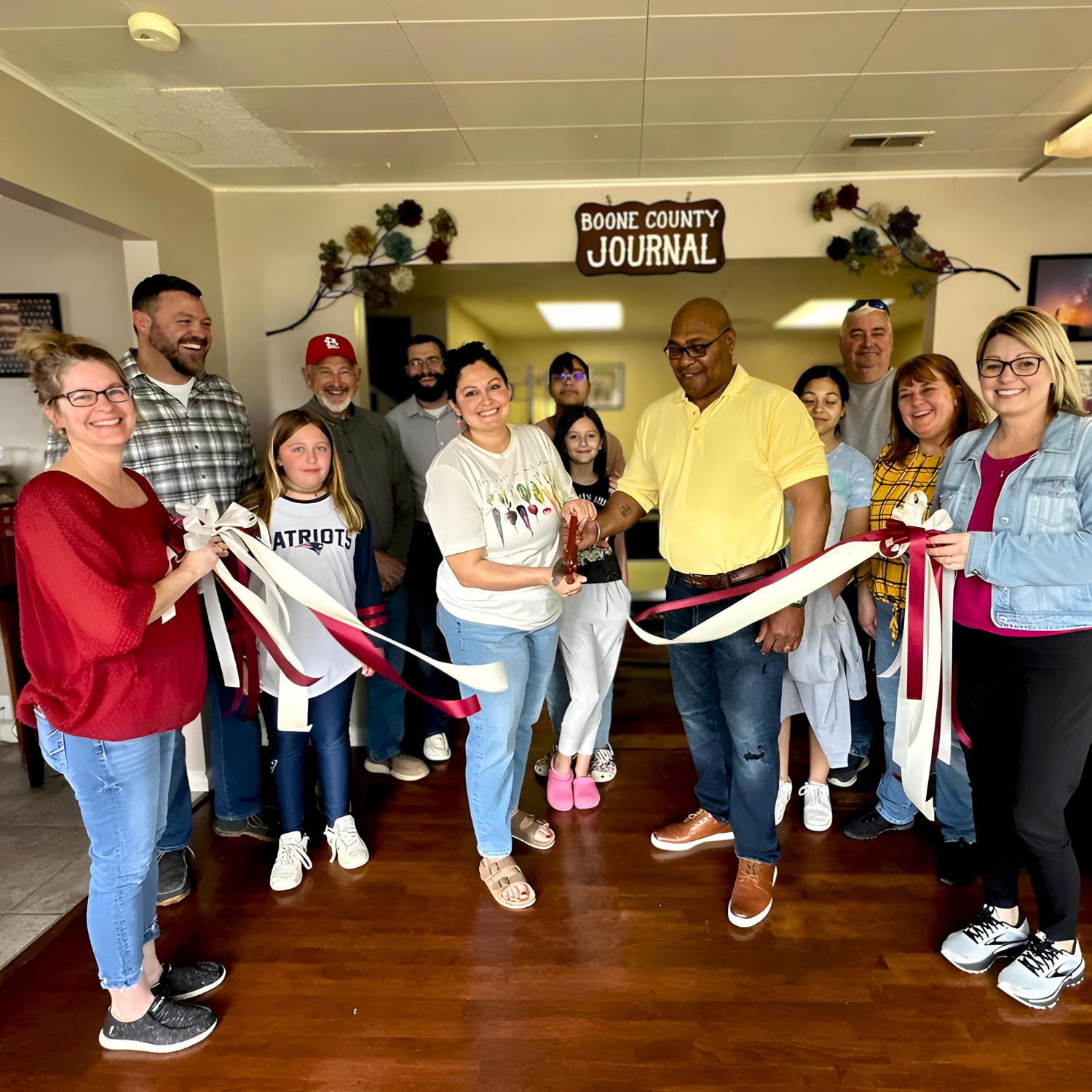 A group of people gathered for a ribbon-cutting event at Boone County Journal, with some holding a ribbon and others smiling.
