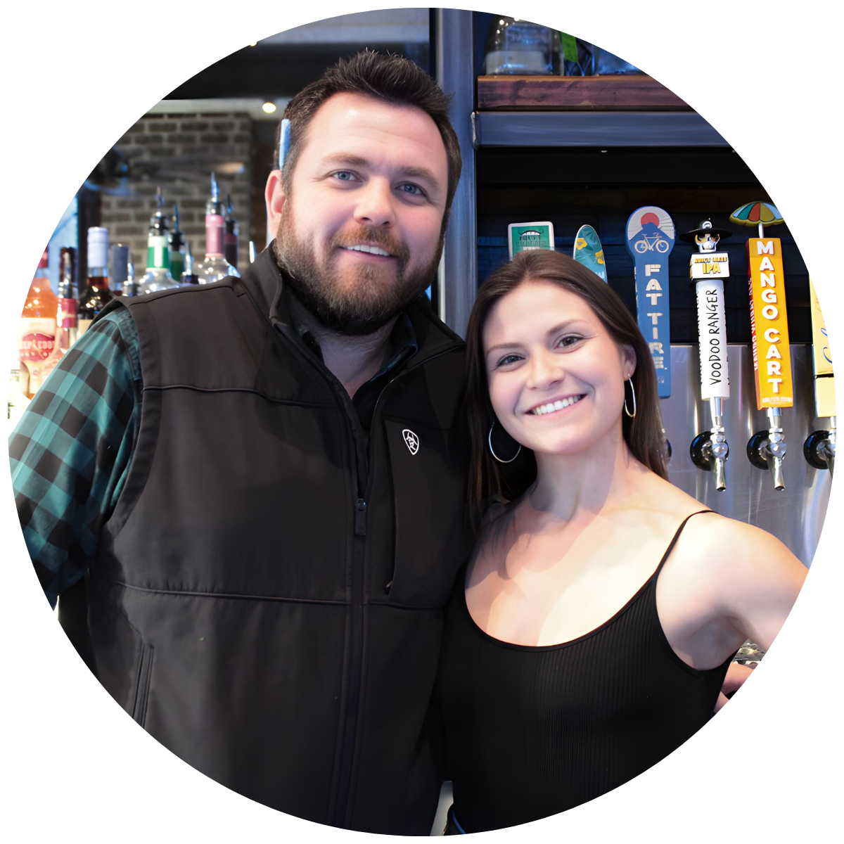 A man and woman smiling together in a bar or restaurant featuring beer taps behind them reading 'Mango Cart' and 'Voodoo Ranger'.
