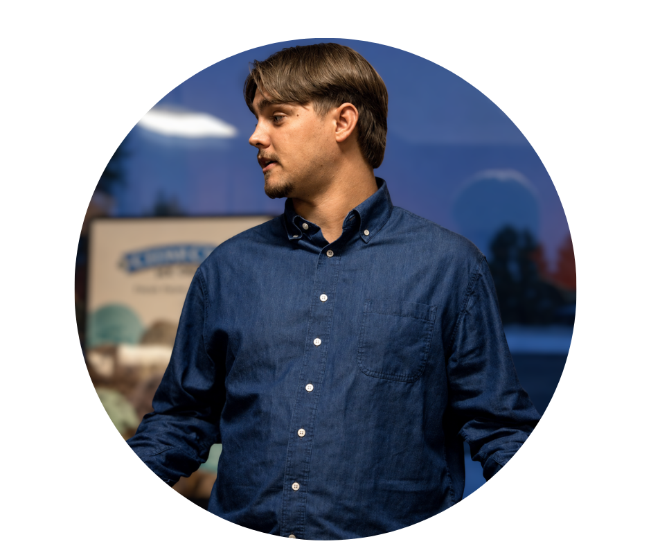 Young man with light brown hair and a goatee wearing a denim button-up shirt, looking to his left outdoors during sunset.