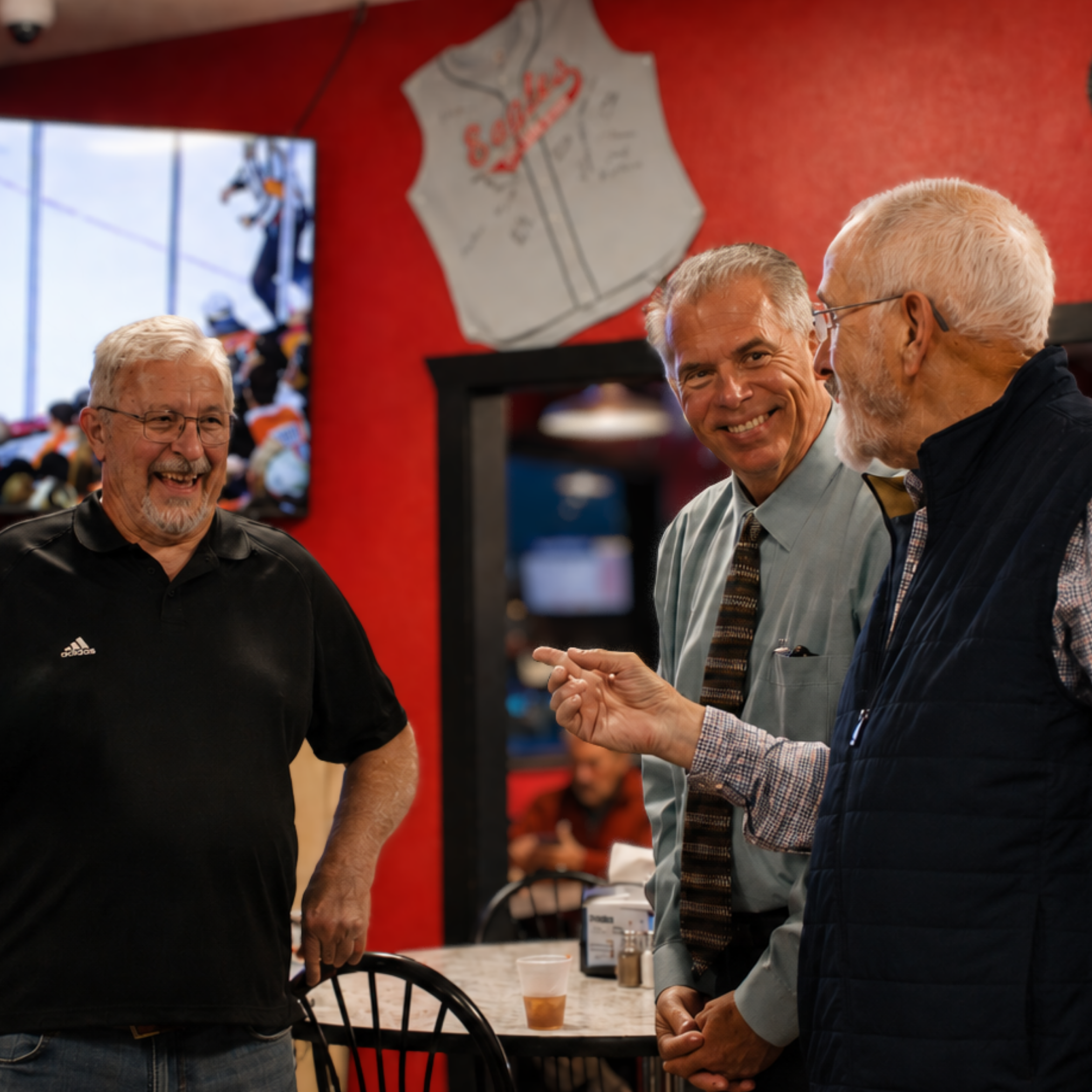 Three men are engaged in conversation in a restaurant. The man on the left is smiling, wearing glasses and a black shirt. The two men on the right are also smiling; one wears a gray shirt while the other has white hair, glasses, and a dark vest. There is a red wall with a hockey jersey and a TV in the background. On the counter, there are a cup of beer, a salt shaker, and other items.