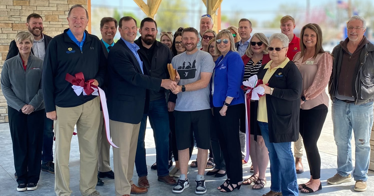 Group of people gathered at a ribbon-cutting ceremony, some holding scissors, celebrating an opening event outdoors under a wooden canopy.