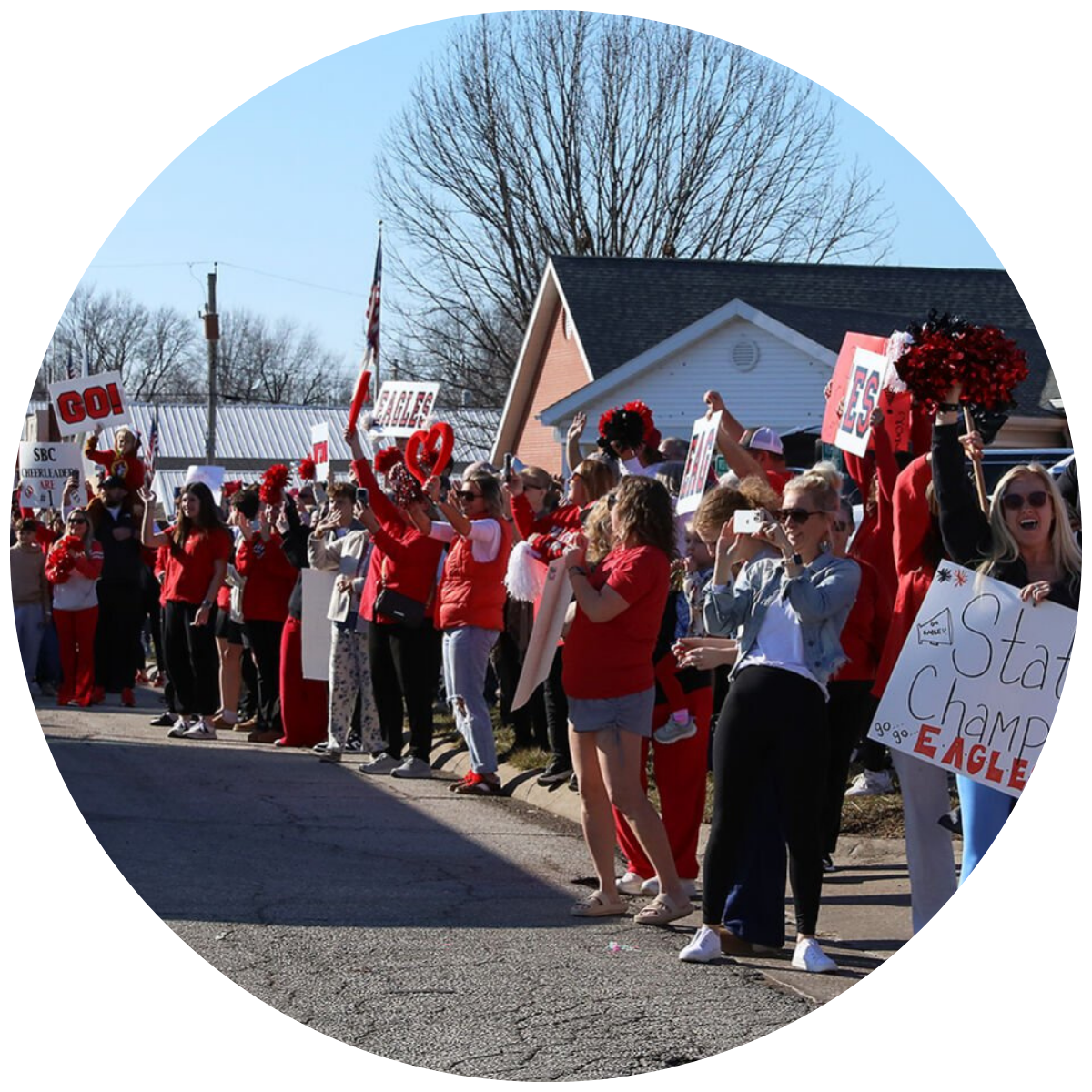 Crowd of people dressed in red and white holding signs and pom-poms, likely at a parade or football game celebration, with some signs reading 'GO!', 'EAGLE', and 'State Champs'.