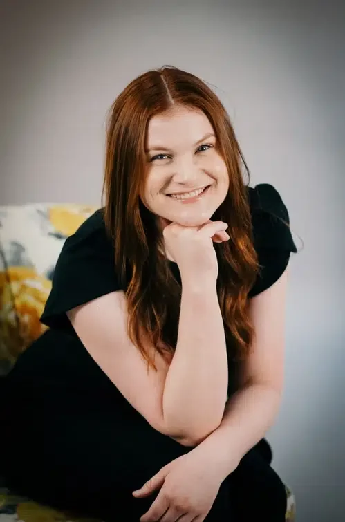 A young woman with long red hair smiling and resting her chin on her hand in a black dress.