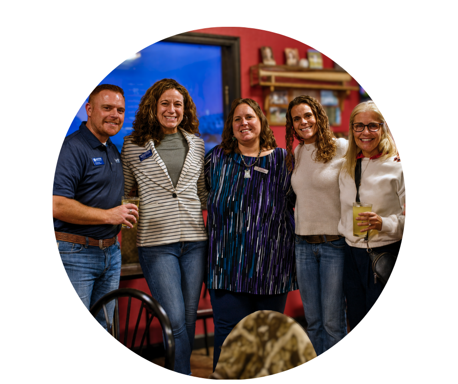 Group of five people smiling and standing together at a social gathering, with drinks in their hands, indoors with red walls and decorative shelving.