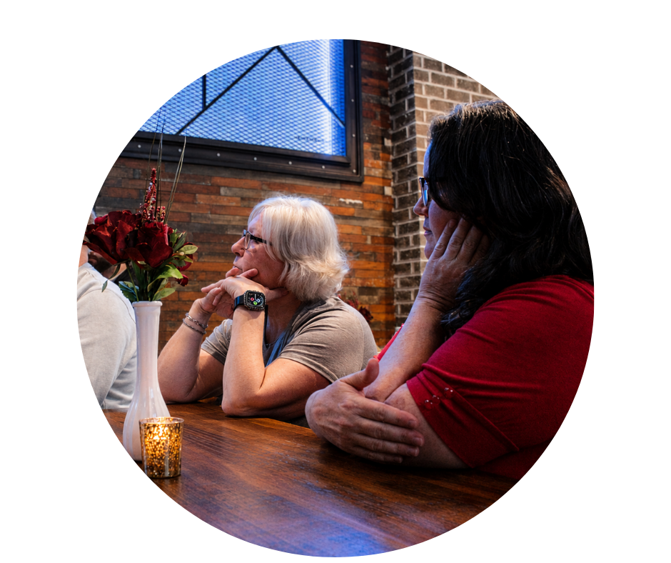 Two women sitting at a wooden table in a cozy restaurant with brick walls. One woman has white hair, glasses, and is wearing a gray shirt; she is looking thoughtfully. The other woman has dark hair, glasses, and is wearing a red shirt; she appears to be resting her head on her hand and is looking away. There is a tall vase with red flowers and a small candle on the table.