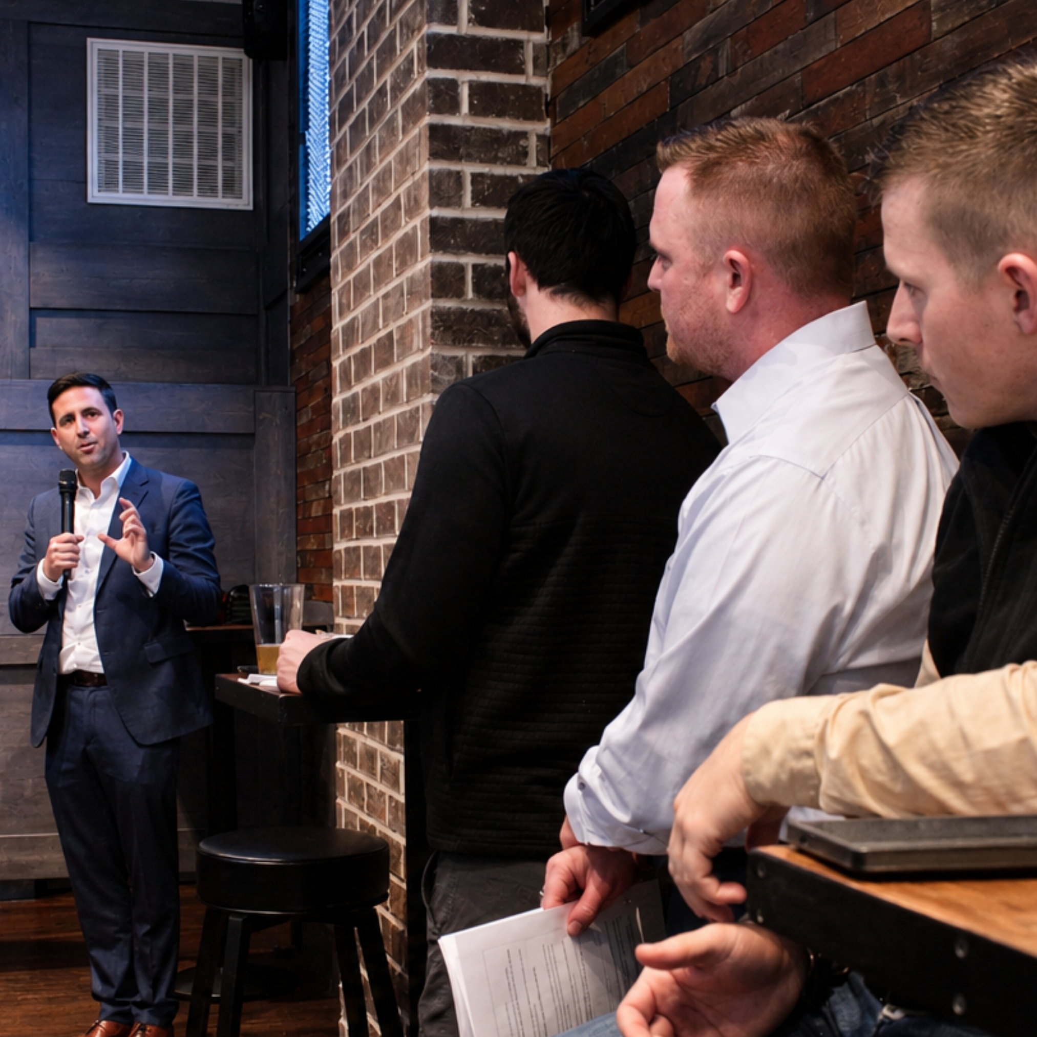 A business seminar or presentation taking place in a rustic-style room with exposed brick and wooden walls, where a man in a suit is speaking into a microphone to an audience.