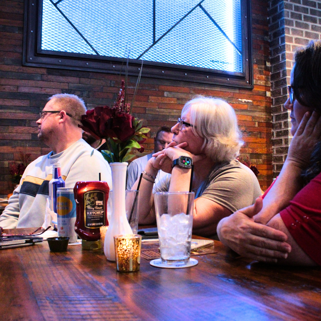 People sitting at a restaurant table with a flower arrangement, ketchup, drink, and condiments, listening attentively, with exposed brick wall and a large window in the background.