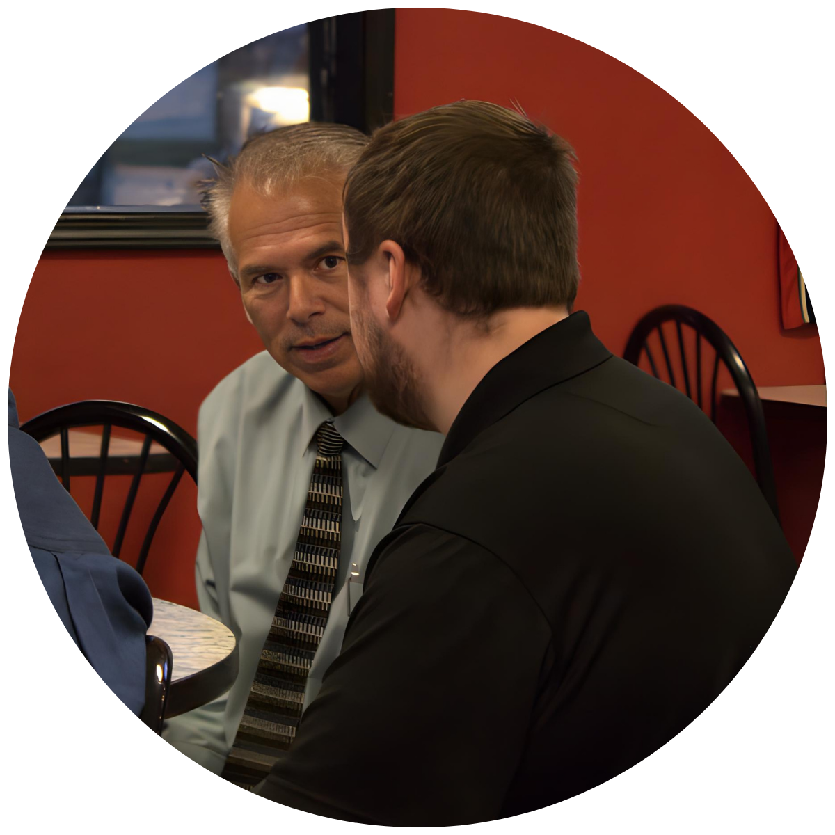 Two men are engaged in a close conversation in a restaurant with red walls and black chairs; one has short gray hair and wears a white shirt with a patterned tie, the other has brown hair and a beard, wearing a black jacket.