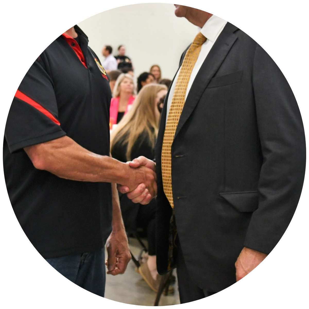 Two men shaking hands at a formal event, with one wearing a black shirt and the other in a dark suit with a yellow tie, in a room with seated people in the background.