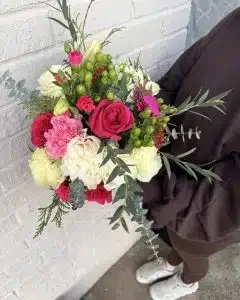 A person holding a colorful flower bouquet with roses, carnations, and greenery, standing against a white brick wall.