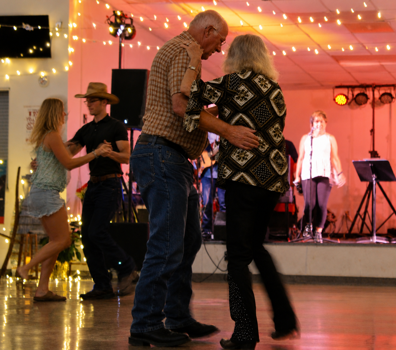 Older man and woman dancing in the foreground at a country-themed dance event, with a band playing on stage behind them. The stage is decorated with string lights and the event has a warm, festive atmosphere.
