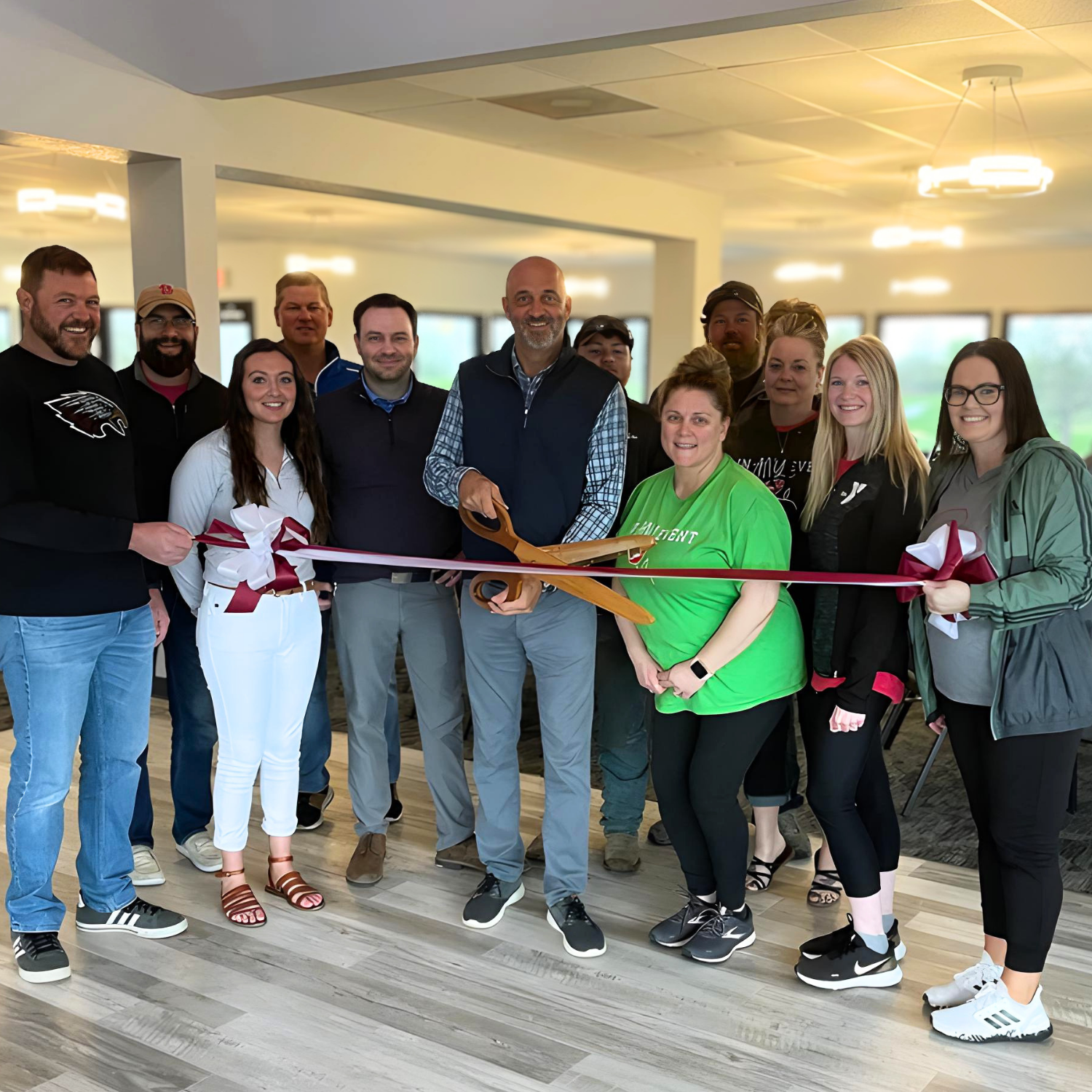 A group of people gathered indoors, participating in a ribbon-cutting ceremony. They are smiling and holding a large pair of gold-colored scissors together.