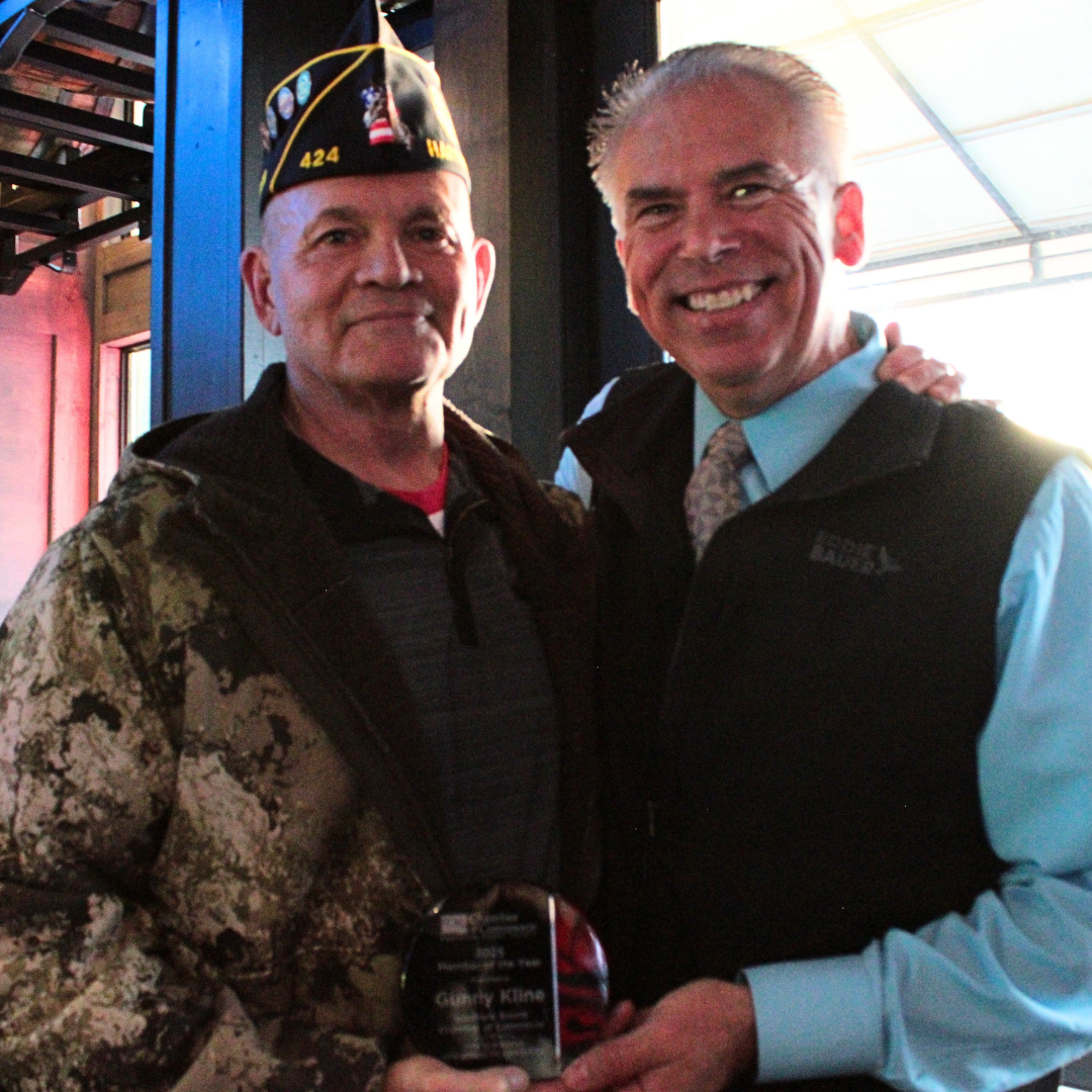 Two men smiling indoors, one dressed in camouflage and a military hat, the other in a dress shirt and vest, holding an award plaque together.