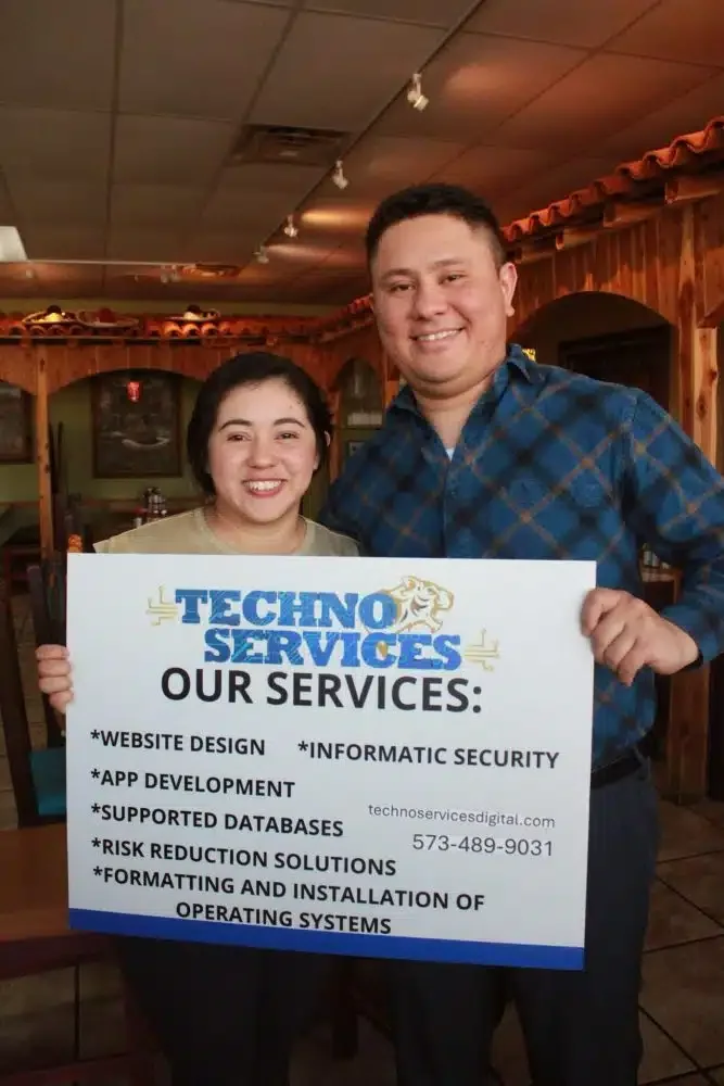 Two people smiling and holding a promotional sign for Techno Services, listing services like website design, app development, and database support, inside a wood-paneled restaurant.