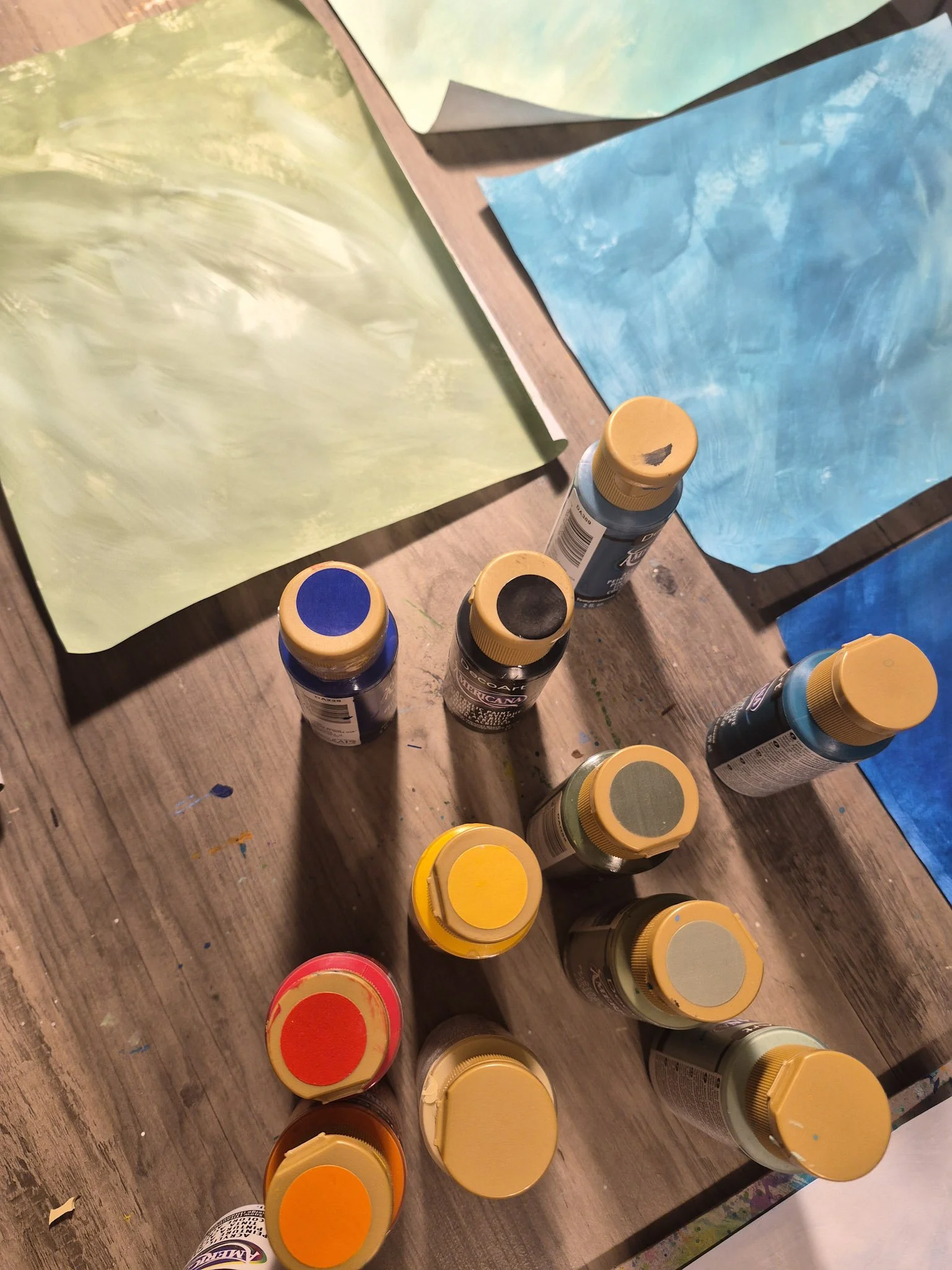 overhead desk shot with a rainbow of paint bottles gathered with hand-painted papers strewn around
