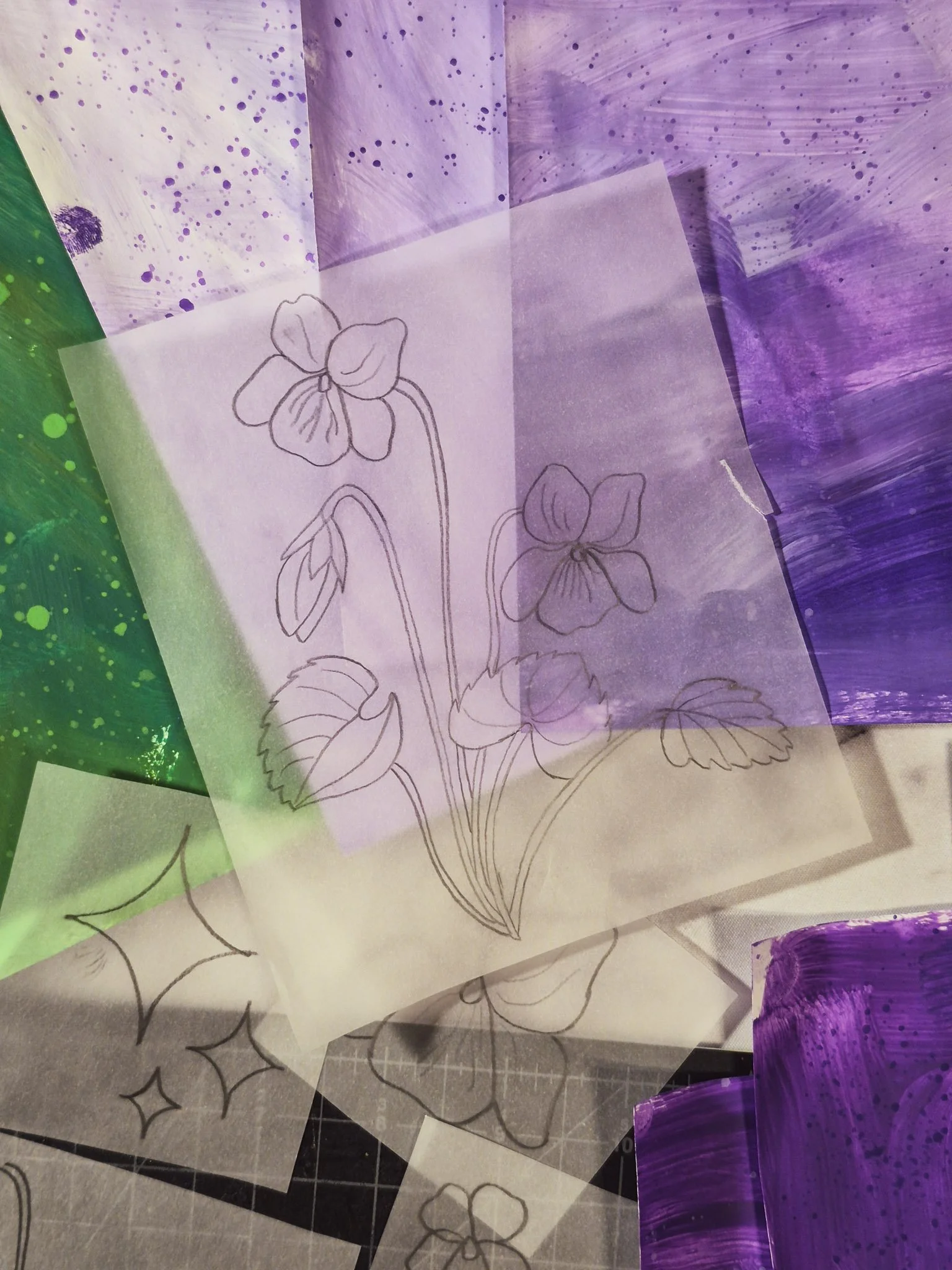 an overhead view of a desk covered in painted collage papers in shades of emerald green and violet with a translucent tracing paper template of a wild violet plant sitting on top.