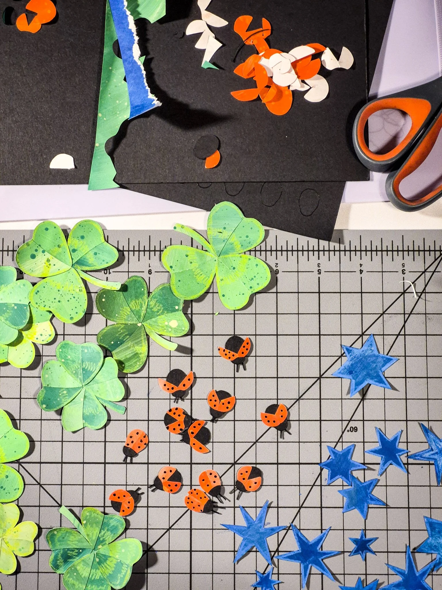 Flat lay of green clover, blue stars, and red ladybug paper collage cutouts arranged on white paper and a cutting mat.