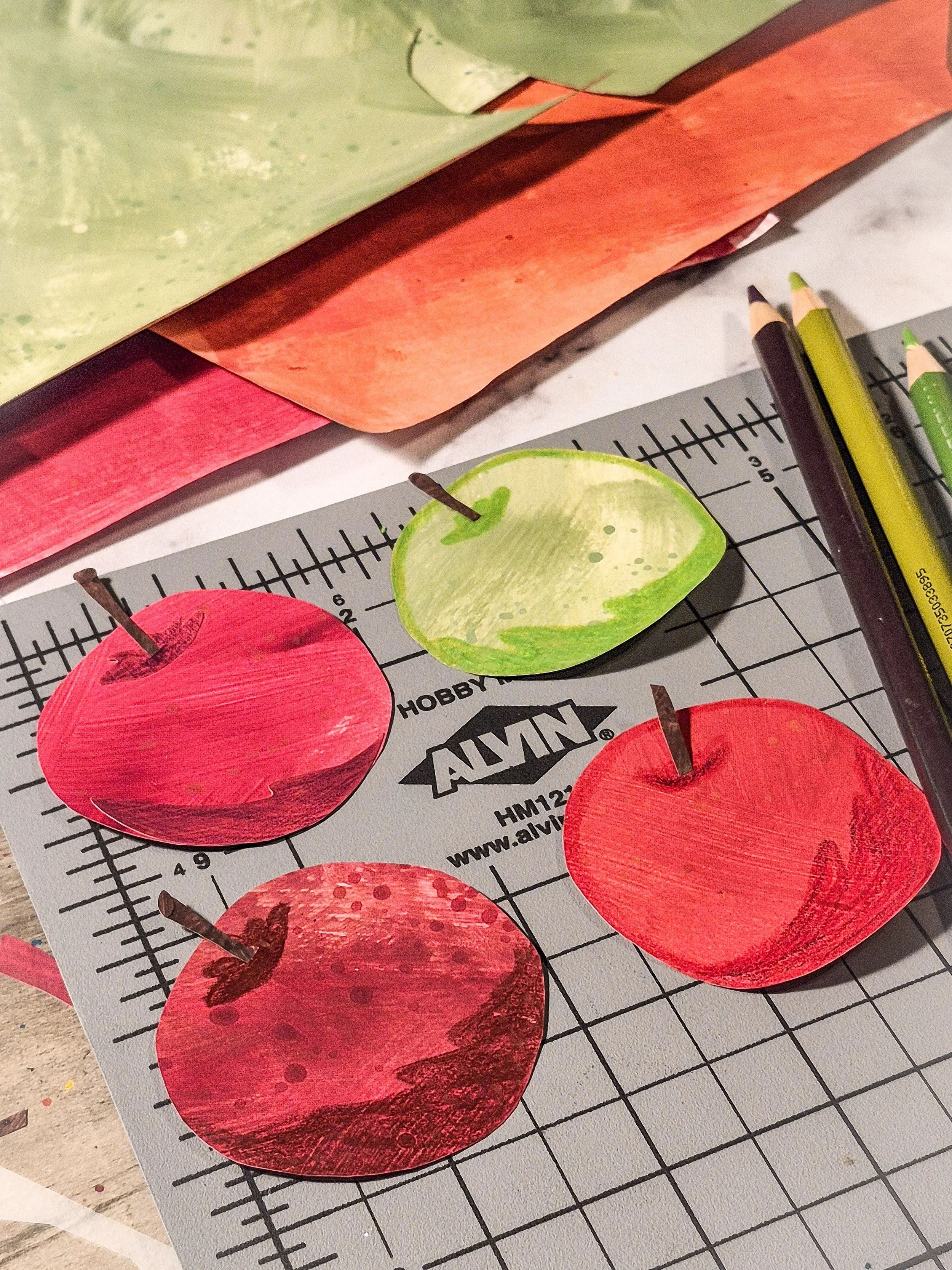 Painted paper apple motifs in shades of red and green on a cutting mat in the studio