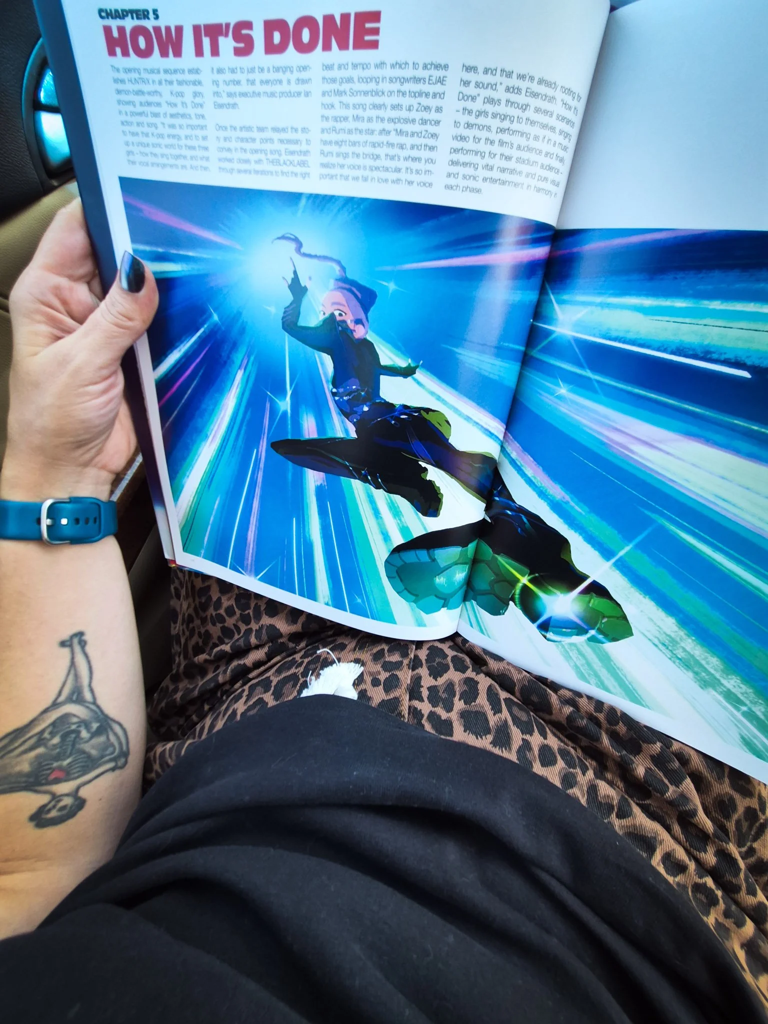 An open illustrated art book showing a vibrant illustration of Rumi, held while sitting in a car.