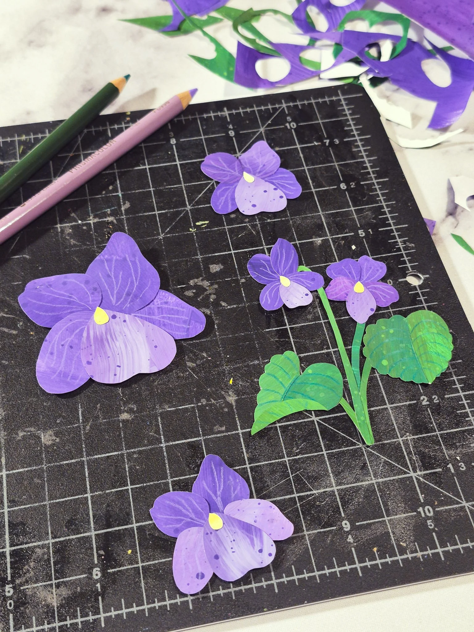 image of a desktop with a black cutting mat and bits of paper scrap scattered around the desk. the mat contains a display of 4 hand-cut violet collages.