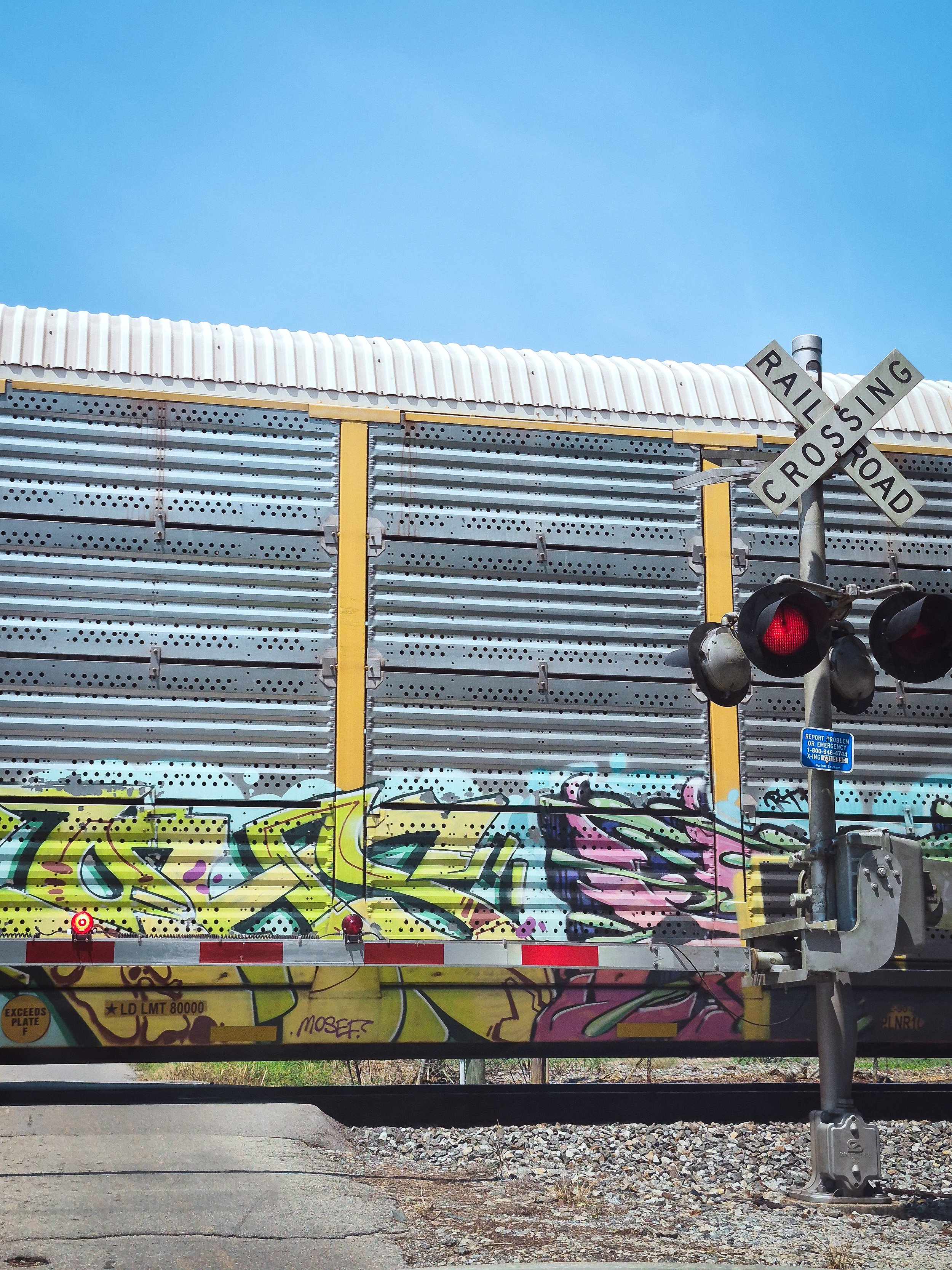Colorful graffiti on a freight train passing a railroad crossing beneath a bright blue summer sky.