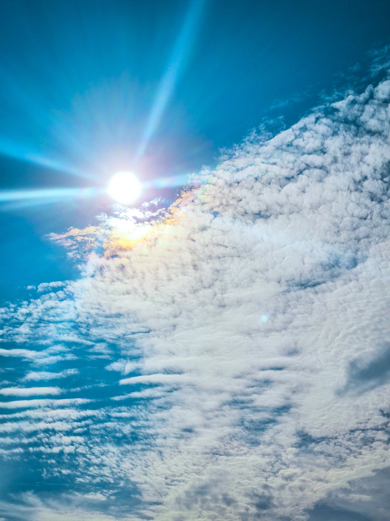 Bright summer sun shining through textured white clouds in a vivid blue sky.
