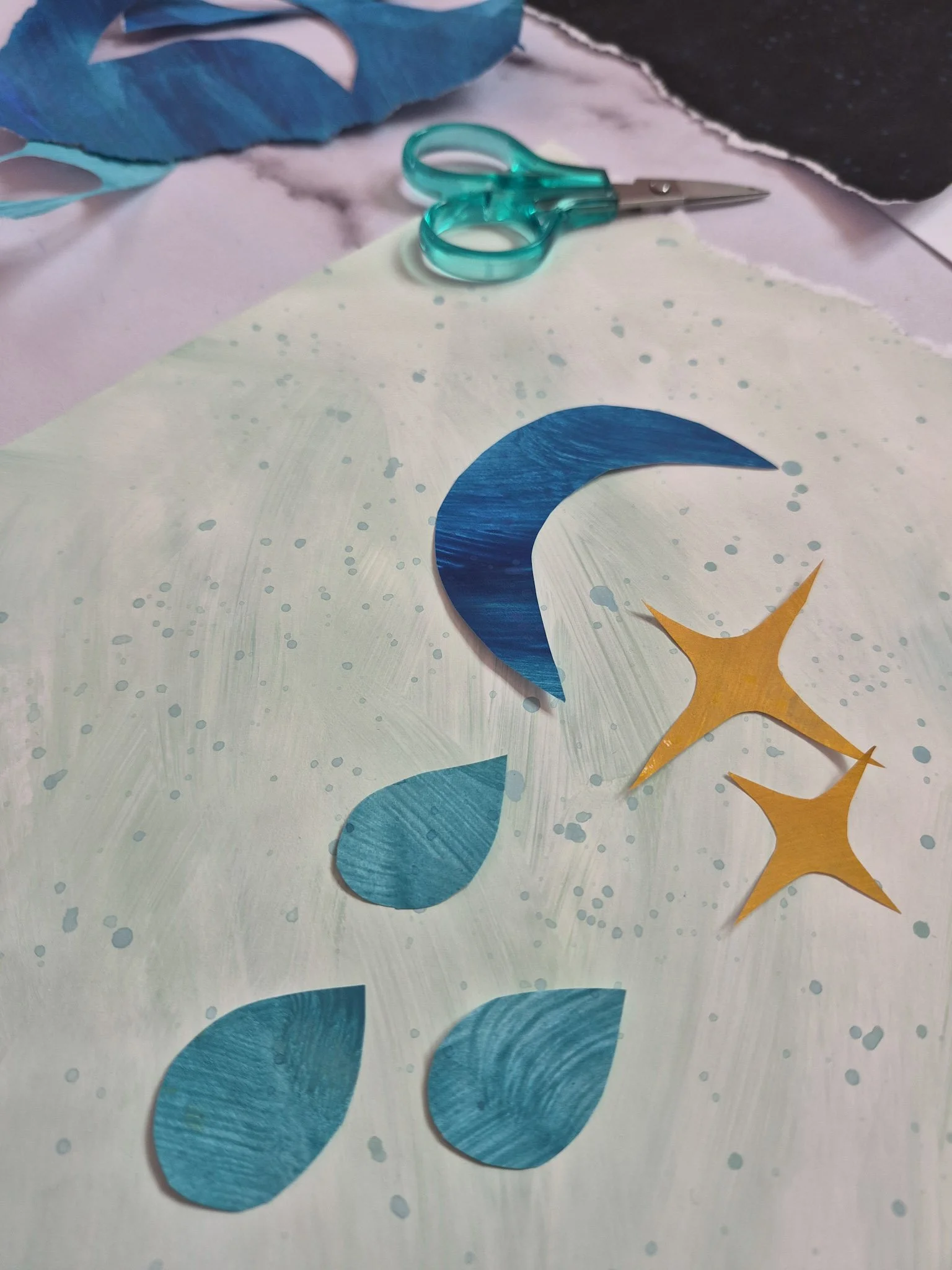 WIP shot of nearly finished collage with blue crescent moon, aqua raindrops, and golden starbursts on a white hand-painted background