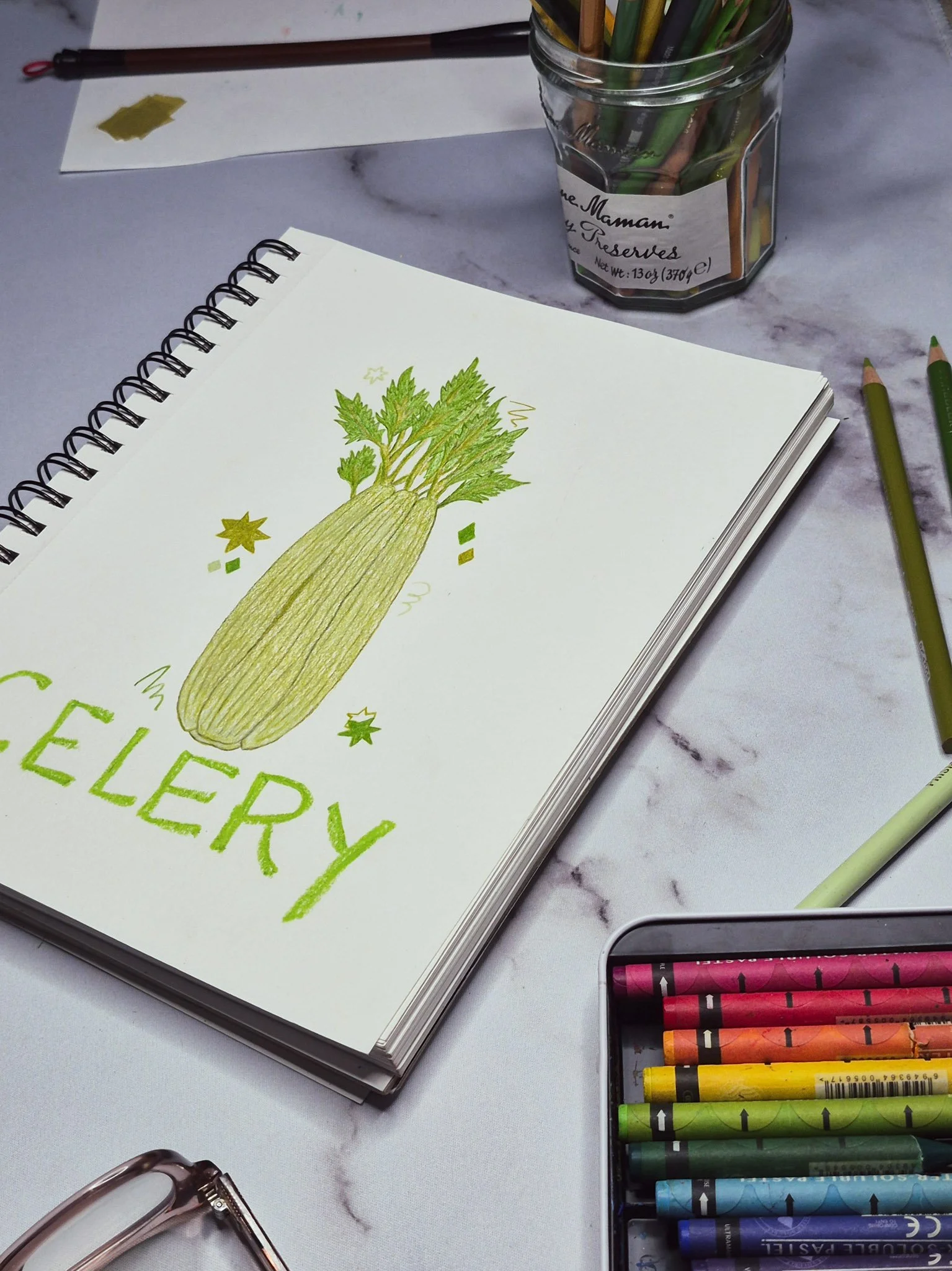 overhead desk shot of completed celery sketch surrounded by water pastels and colored pencils on a marble background